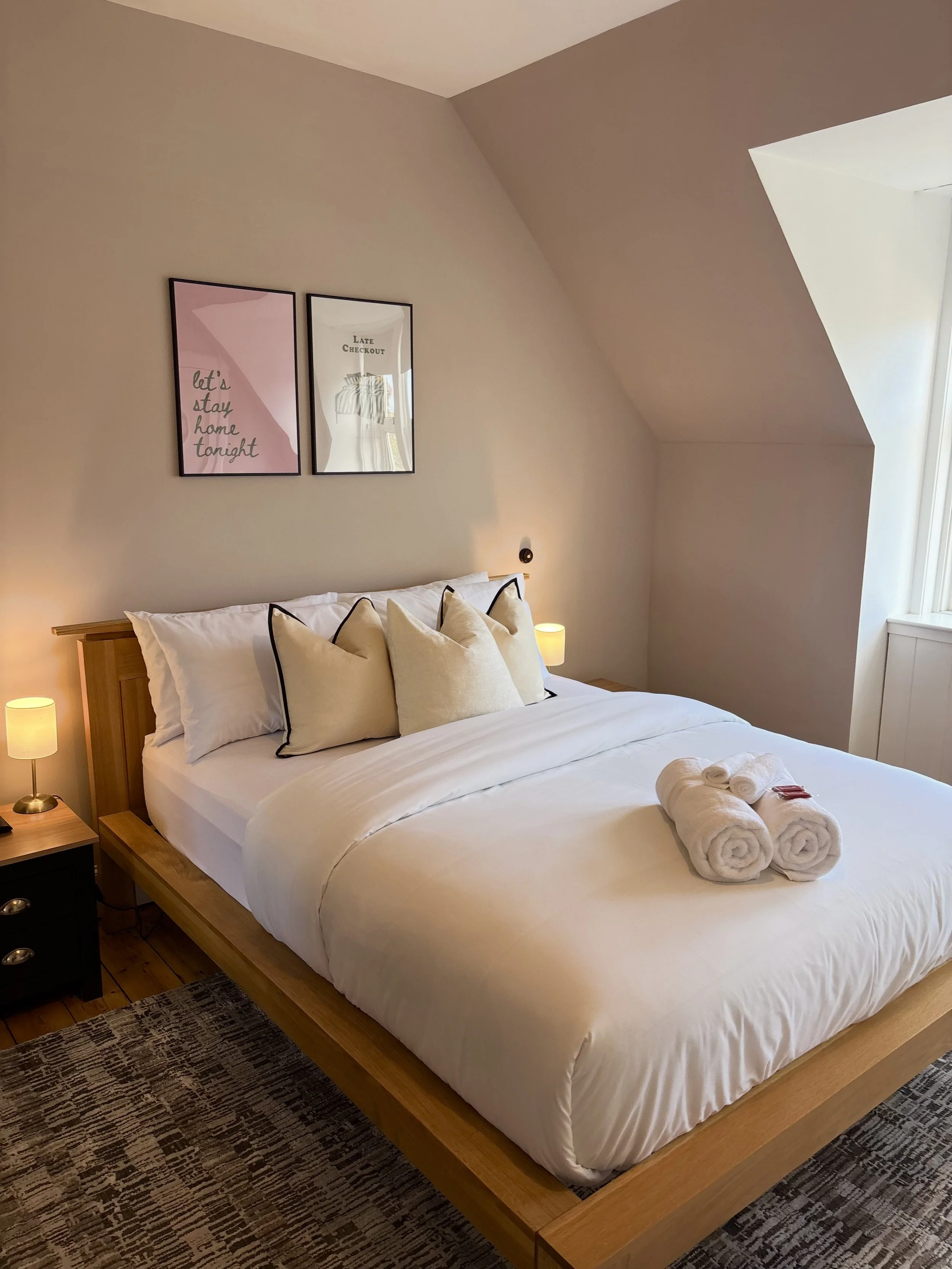 A neatly made bed with white linen and five decorative pillows, two rolled towels, a nightstand with a small lamp, wall art above the bed, and a window with natural light in the corner of a cozy bedroom with sloped ceiling.