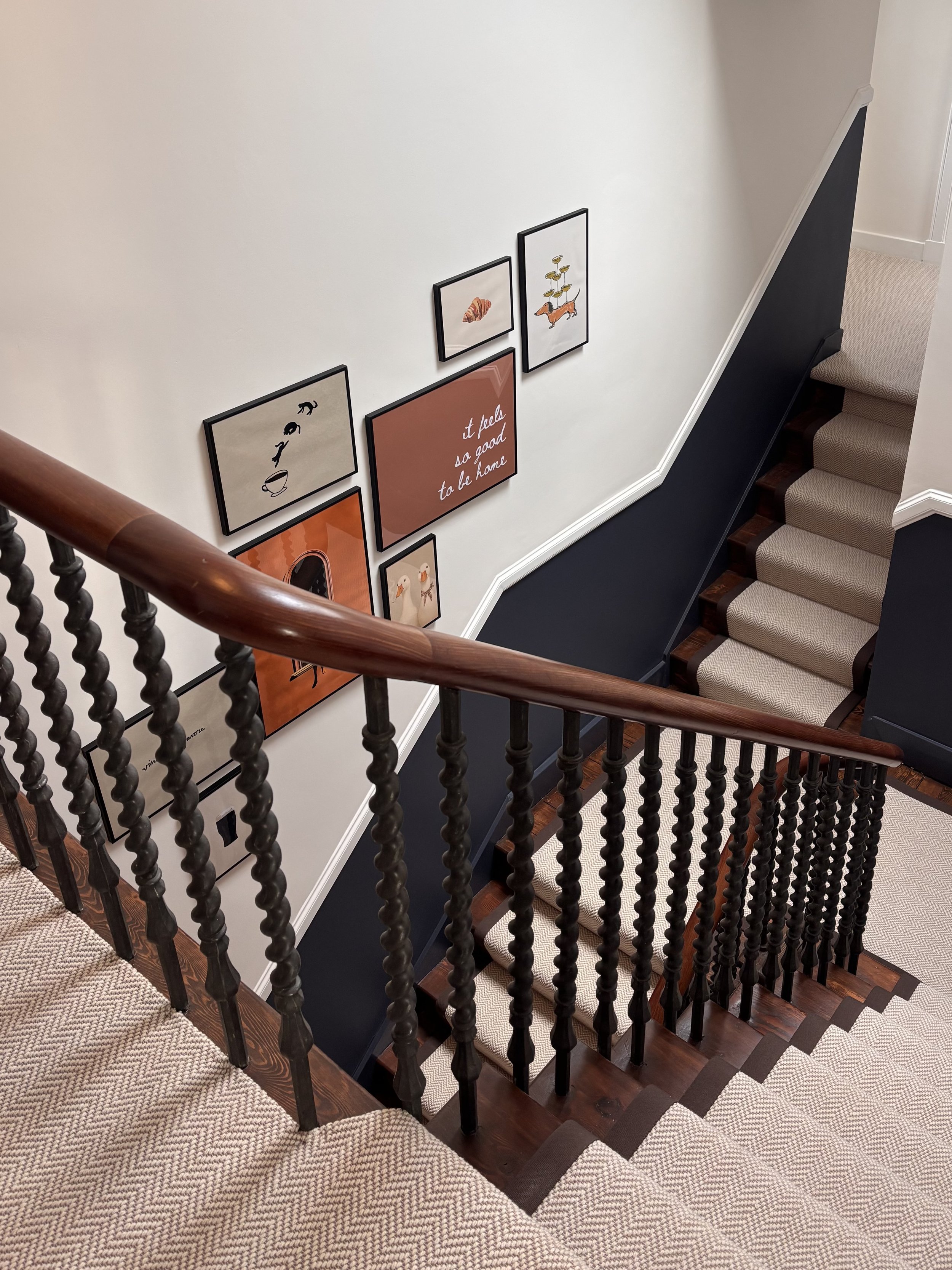 Staircase viewed from the top, with beige carpeted stairs, black side panel, wooden handrail, and wall decor with framed artwork and text.