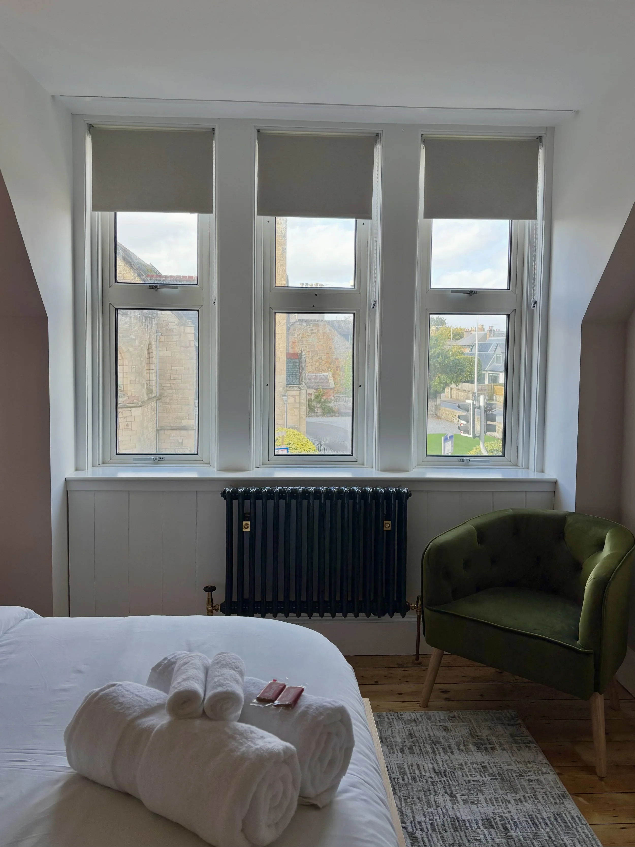 A hotel or guest room with a large window, a bed with towels and toiletries, a green armchair, a radiator, and a patterned rug.