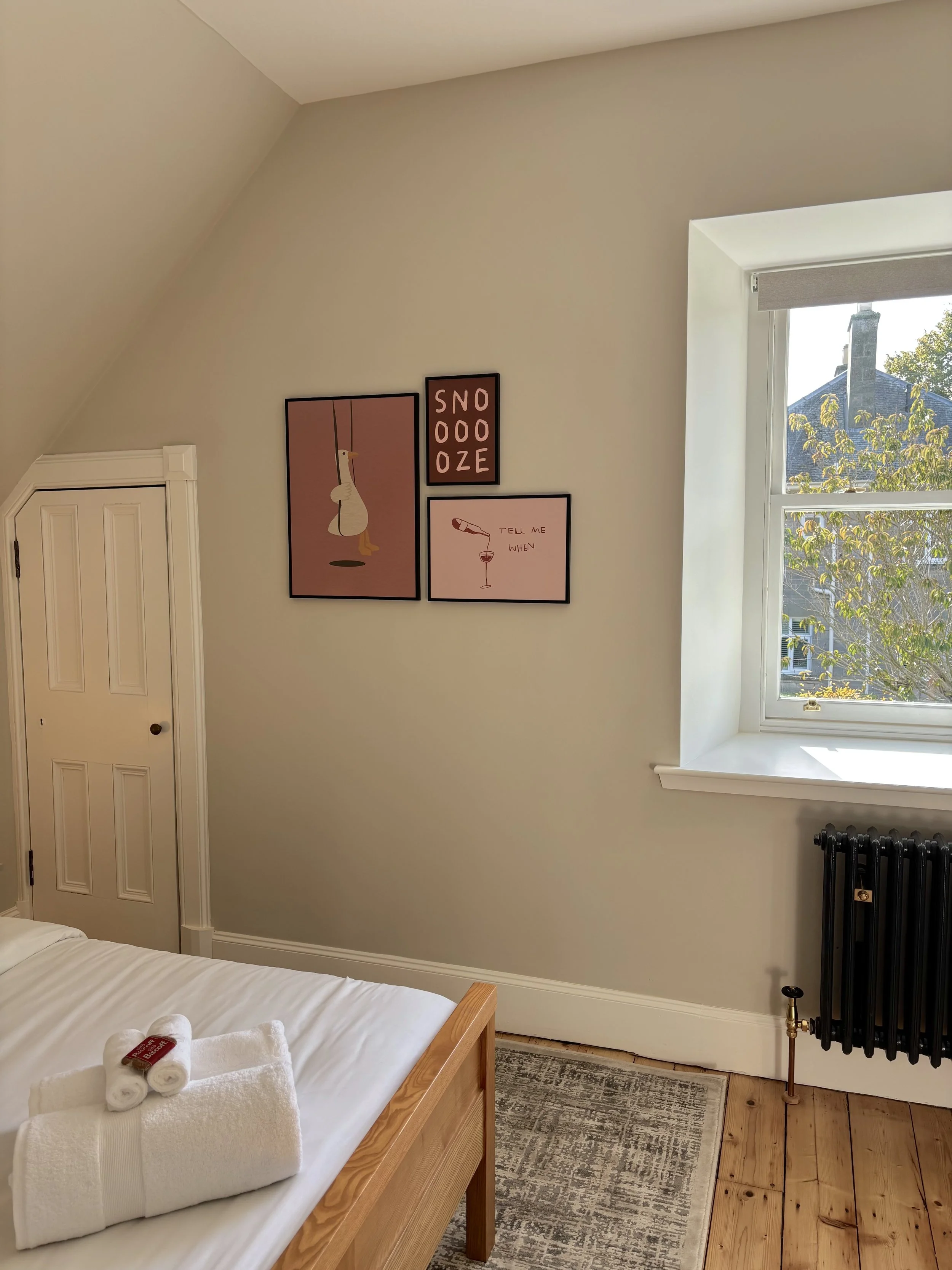 A bedroom with a bed, towels, and toiletries on it, four framed art prints on the wall, a window with a view of trees and rooftops outside, and a radiator near the window.