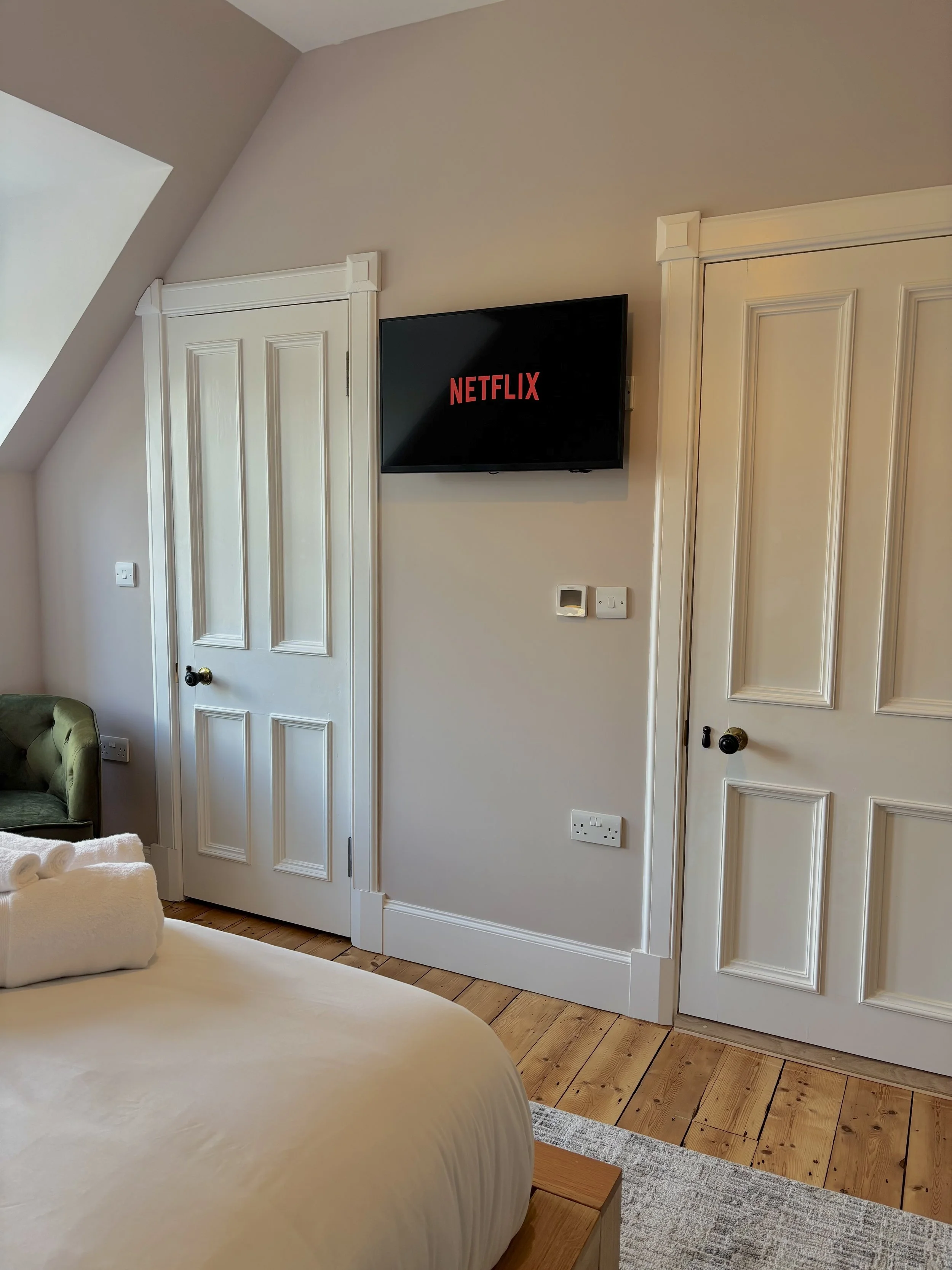 Bedroom with beige walls and wooden floor. A bed with cream bedding is in the foreground. Two white paneled closet doors are on the wall, with a small flat-screen TV showing the Netflix logo mounted between them. There is a green armchair in the corn
