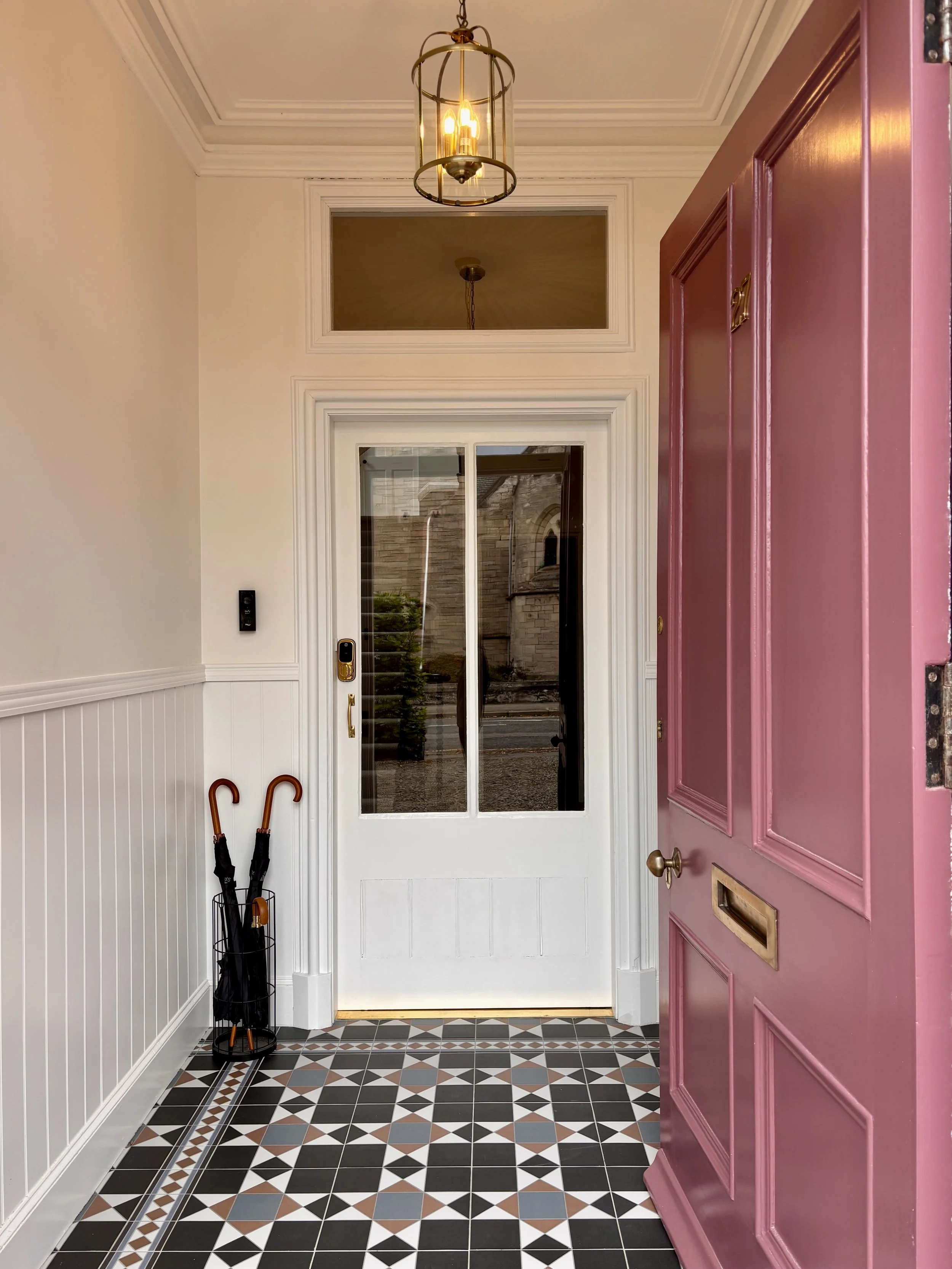 Entryway with a pink door, black, gray, white, and terracotta patterned tile floor, white walls with wainscoting, and a glass sliding door leading outside. There is a black umbrella stand with umbrellas and a hanging lantern-style ceiling light.
