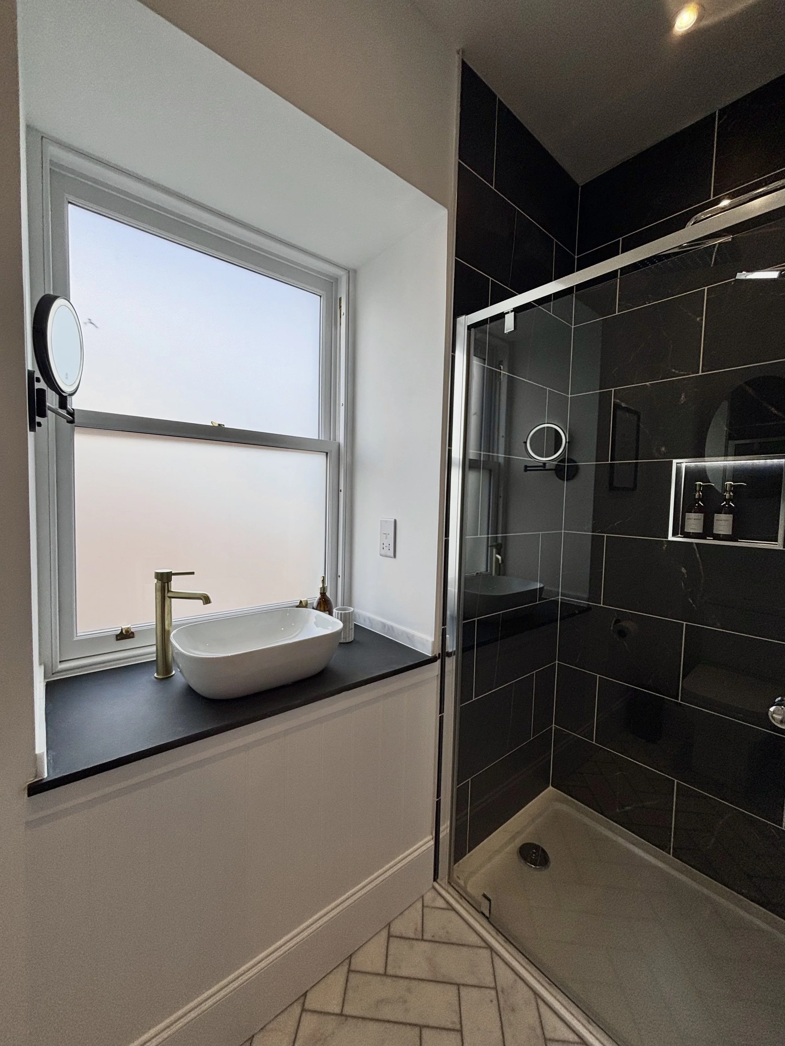 A modern bathroom with a white sink and black countertop, a window above the sink, a black tiled shower area with a glass door, and beige floor tiles.