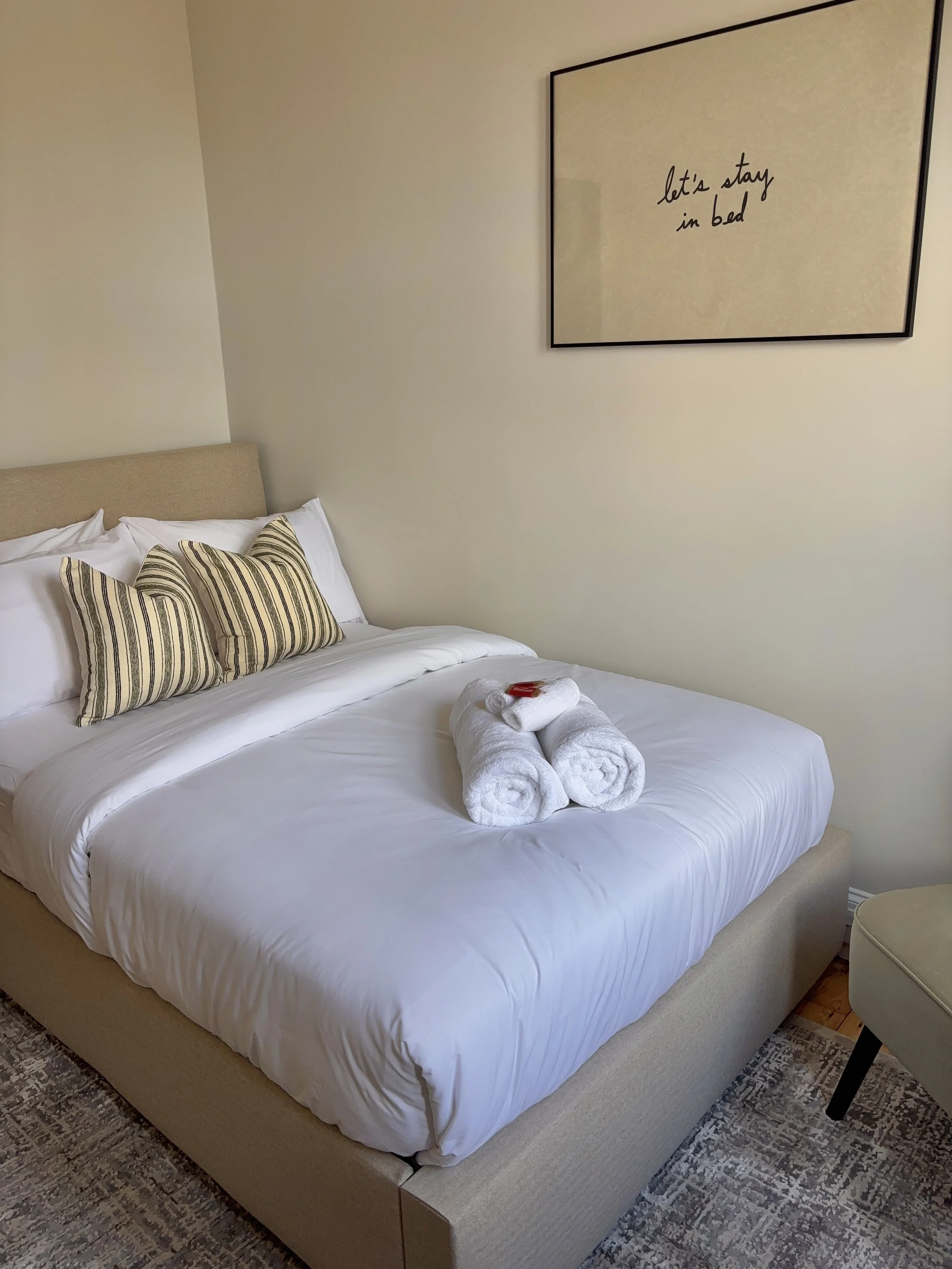 A neatly made bed with white sheets and two rolled white towels on top. There are three striped pillows resting against a beige headboard. A framed sign on the wall reads 'let's stay in bed'.