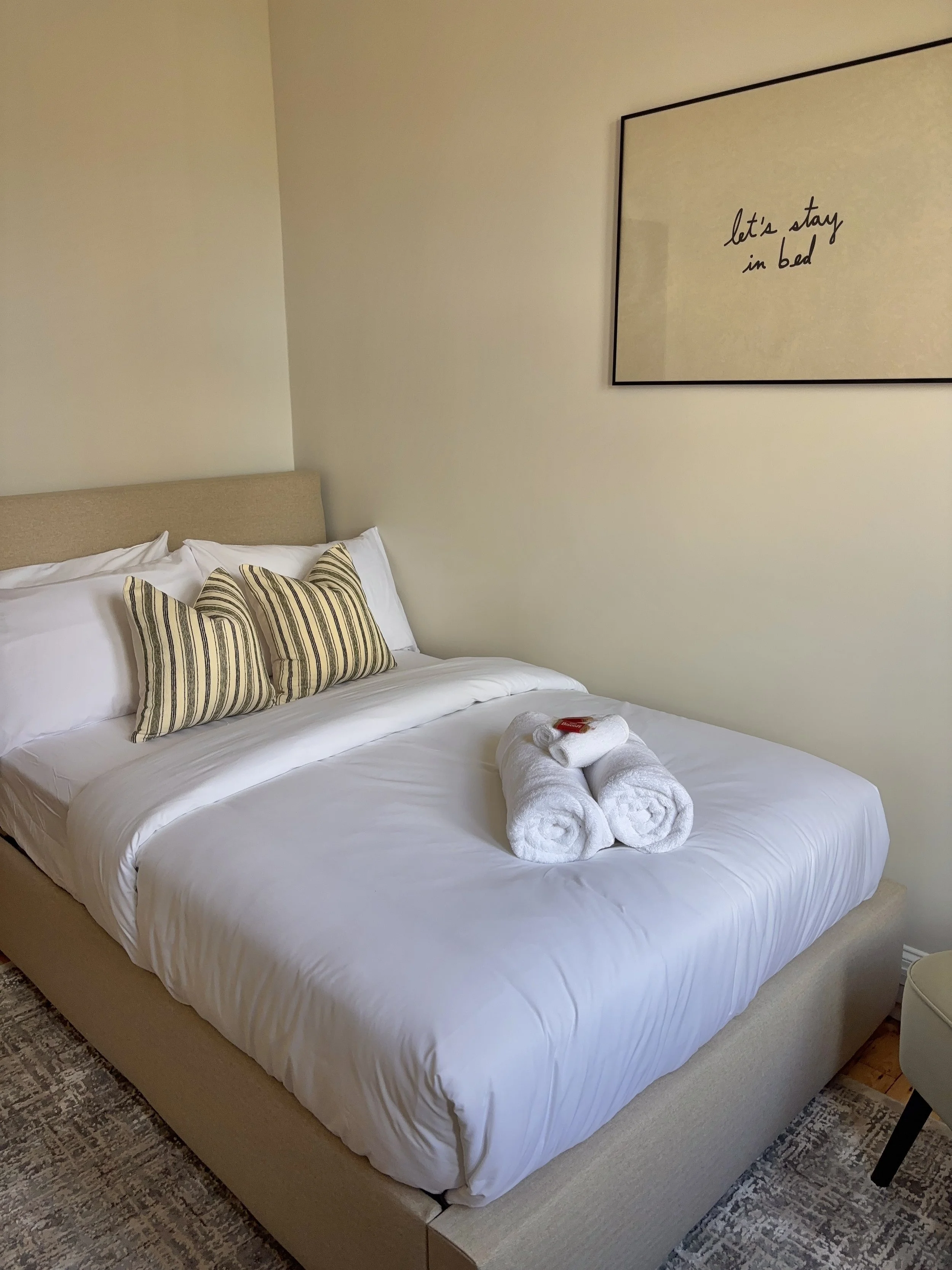 A neatly made bed with white sheets and pillows, two rolled white towels with a small red soap on top placed on the bed, beige headboard, wall art with the phrase 'let's stay in bed', and a dark chair with a white cushion beside the bed.