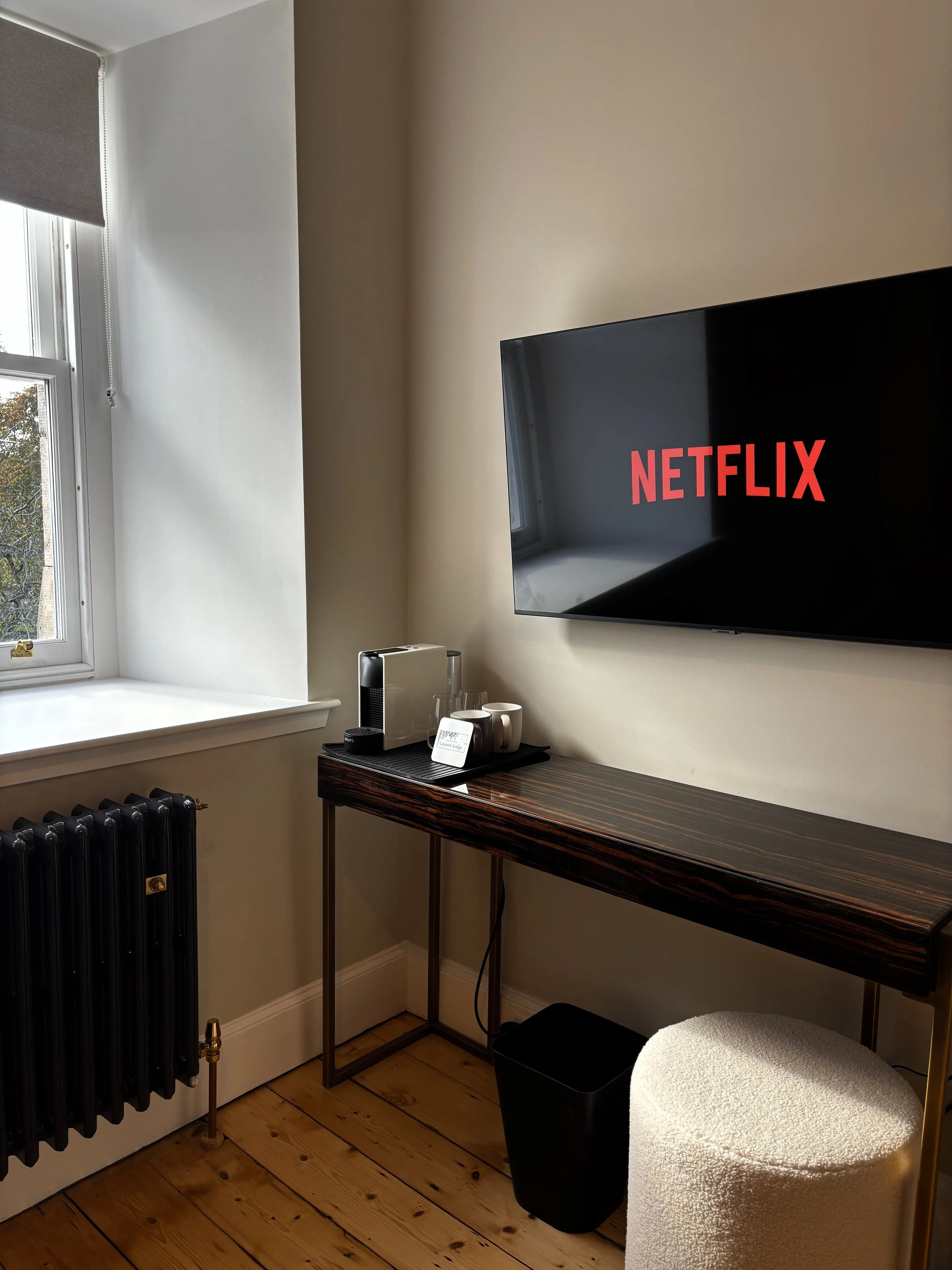 A room with a window, a wall-mounted TV displaying Netflix, a small desk with a coffee machine and mugs, a black trash bin, a round stool, a radiator, and a wooden floor.