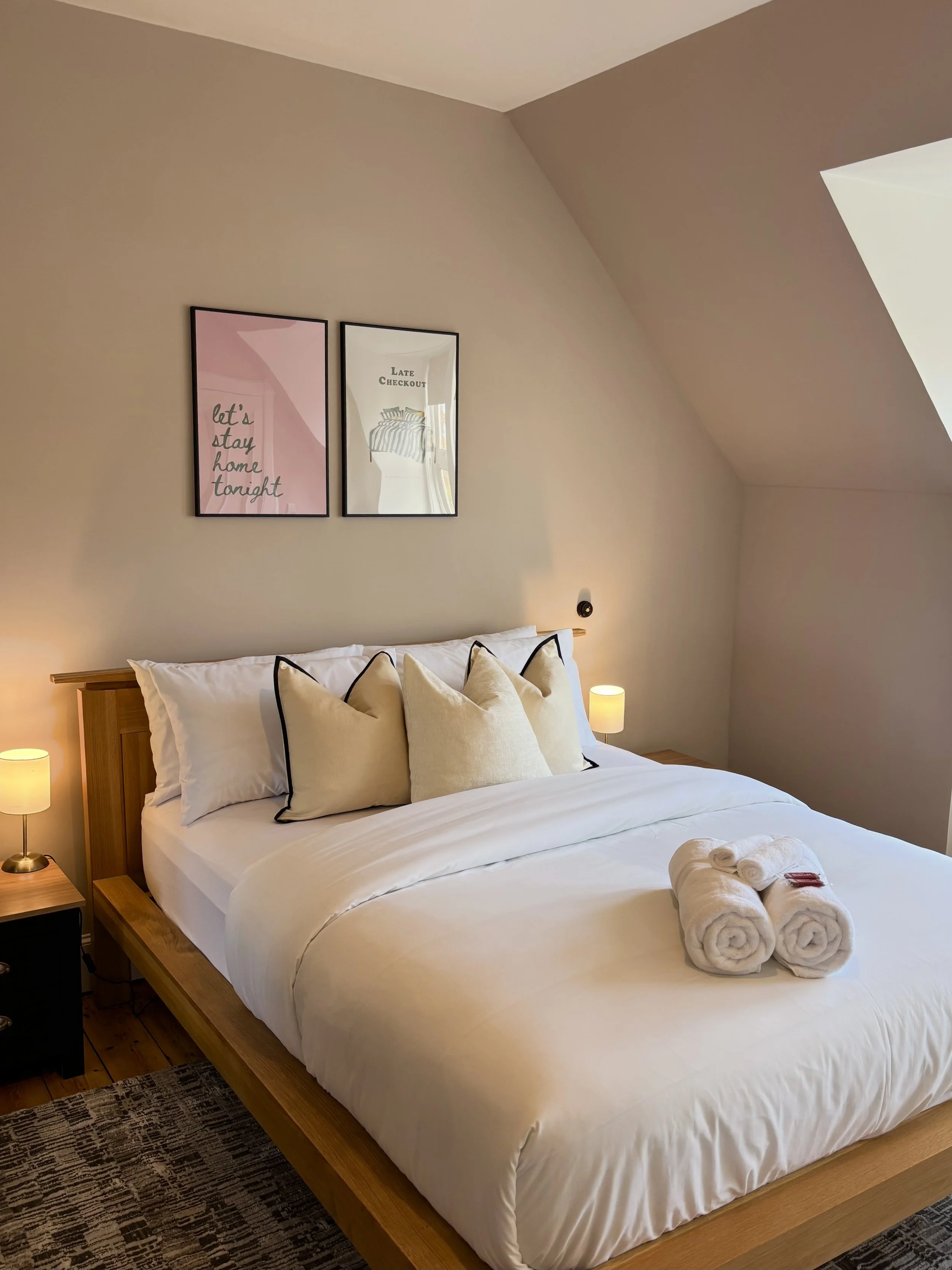 A neatly made bed with white linens and multiple decorative pillows, flanked by two small nightstands with lamps, in a well-lit bedroom with beige walls and framed artwork above the bed. Towels and toiletries are placed on the bed.