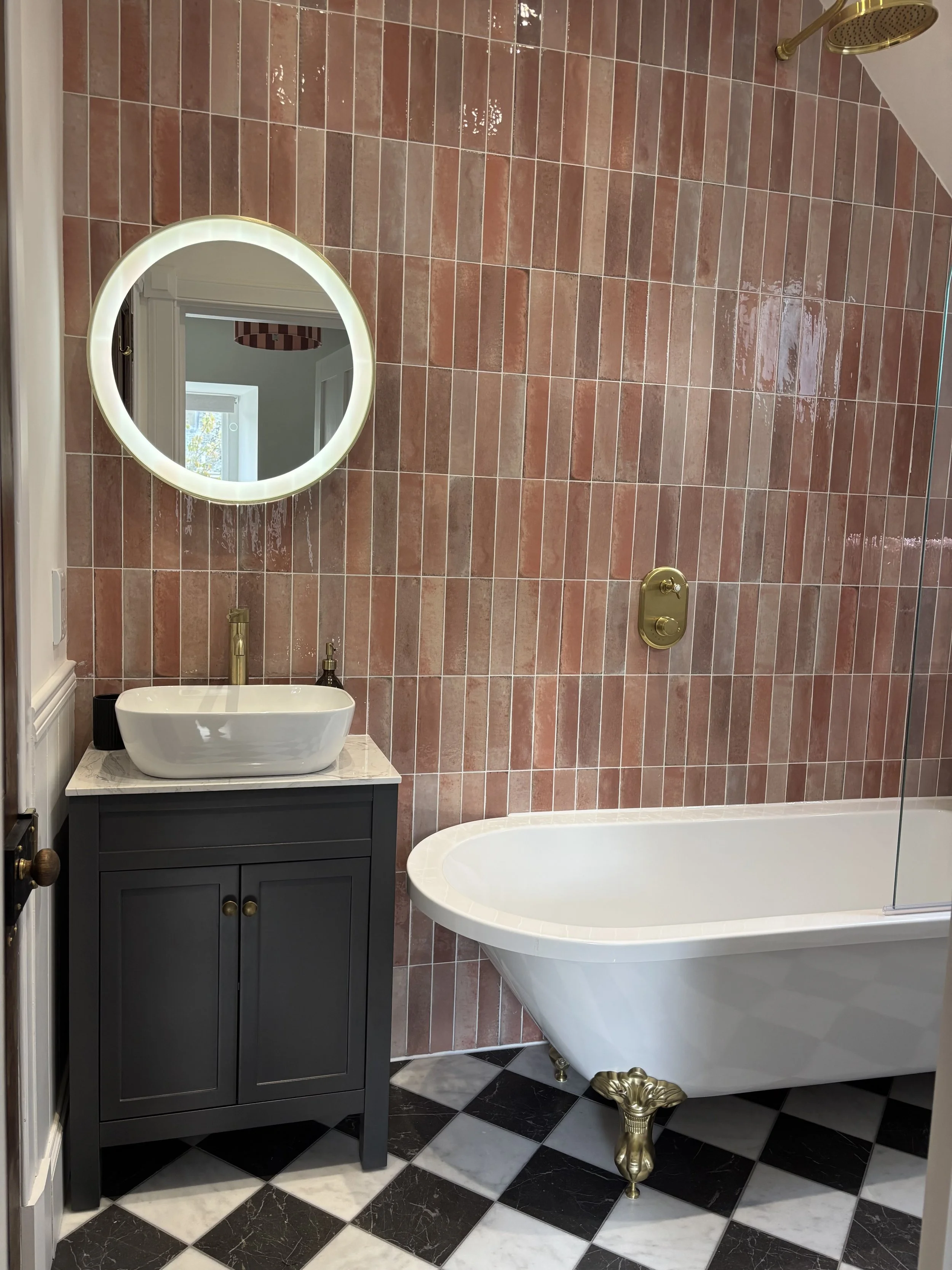Bathroom with pink and white tiled wall, round illuminated mirror, white vessel sink on a dark gray cabinet, gold fixtures, and a clawfoot bathtub with gold clawfoot.