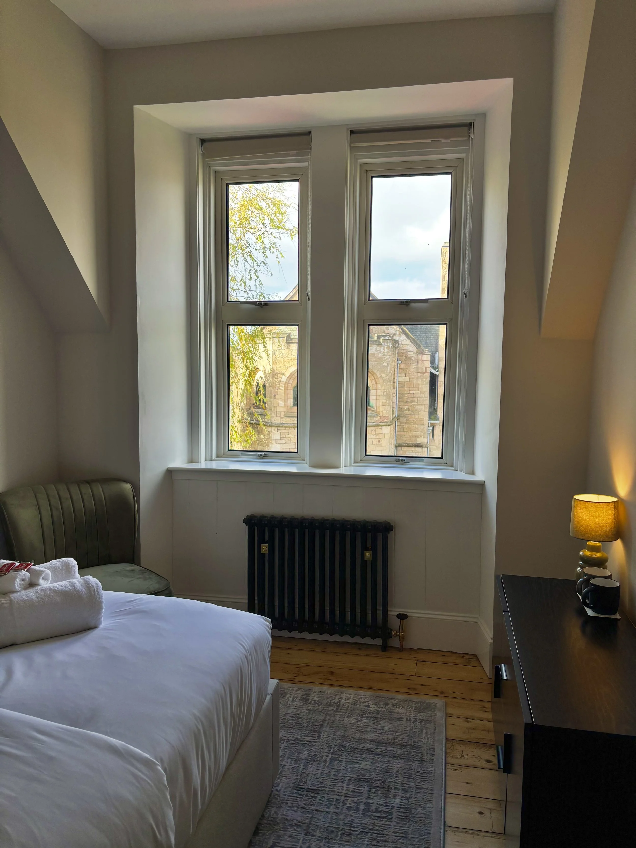 A hotel room with a large window, a bed with white linens and a rolled towel, a small carpet, a dark furniture piece with a lamp, and a radiator below the window. Outside the window, a tree and a stone building are visible.