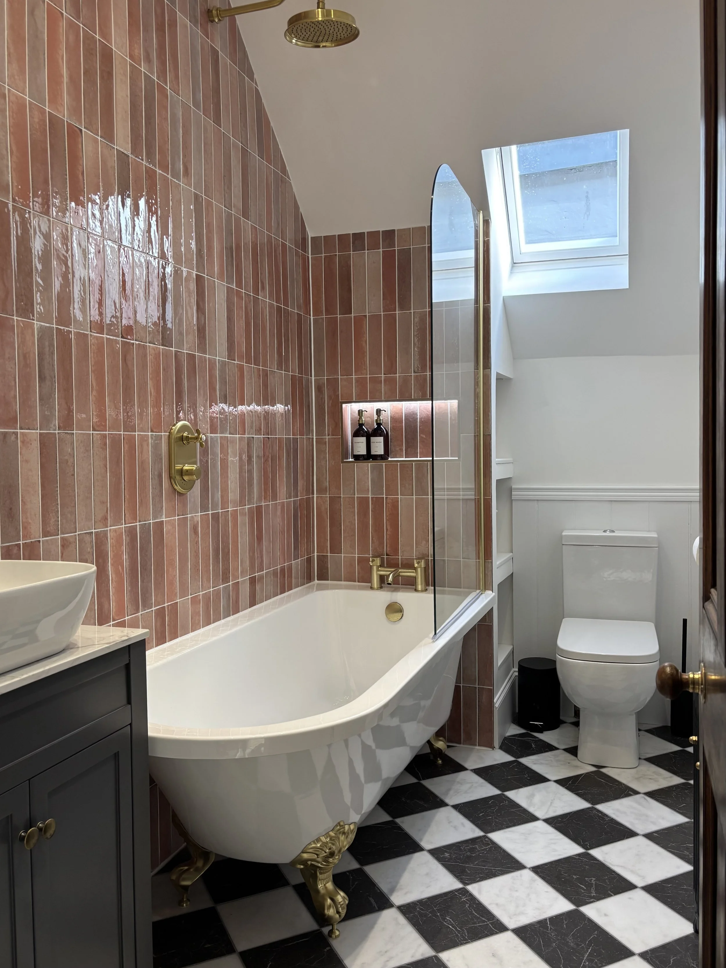 Bathroom with a vintage clawfoot bathtub, brass fixtures, black and white checkered floor, gray vanity, white toilet, built-in shelf with bottles, window with skylight, and a brass showerhead.