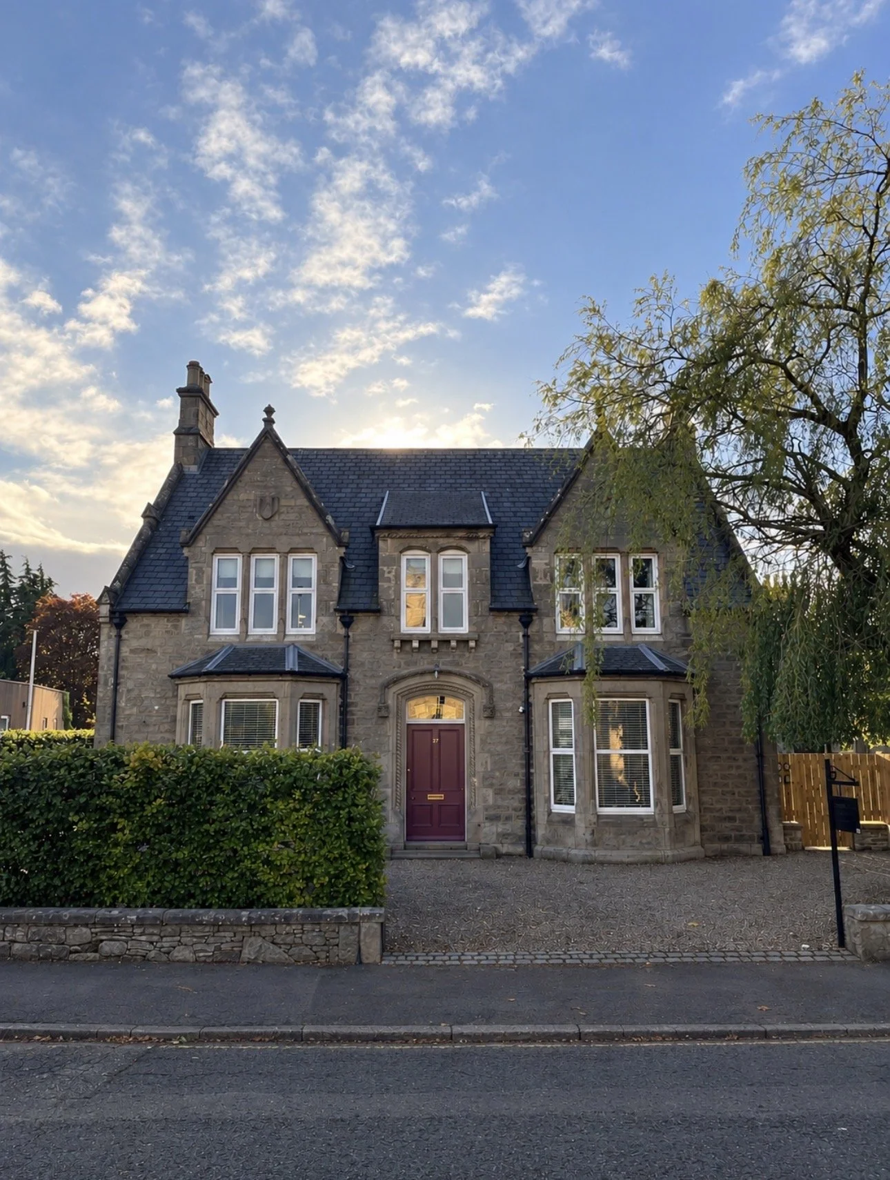 A large stone house with a pointed roof, multiple white-framed windows, and a red front door, surrounded by greenery and under a partly cloudy sky.