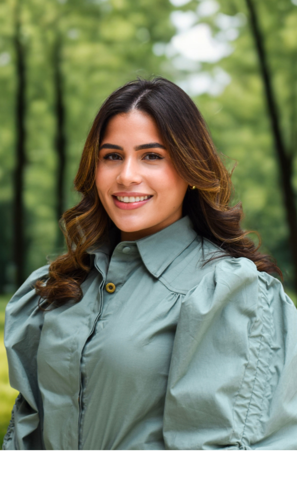 A woman with long wavy brown hair smiling and wearing a green buttoned shirt, standing in front of a light-colored wall and a bronze sculpture.