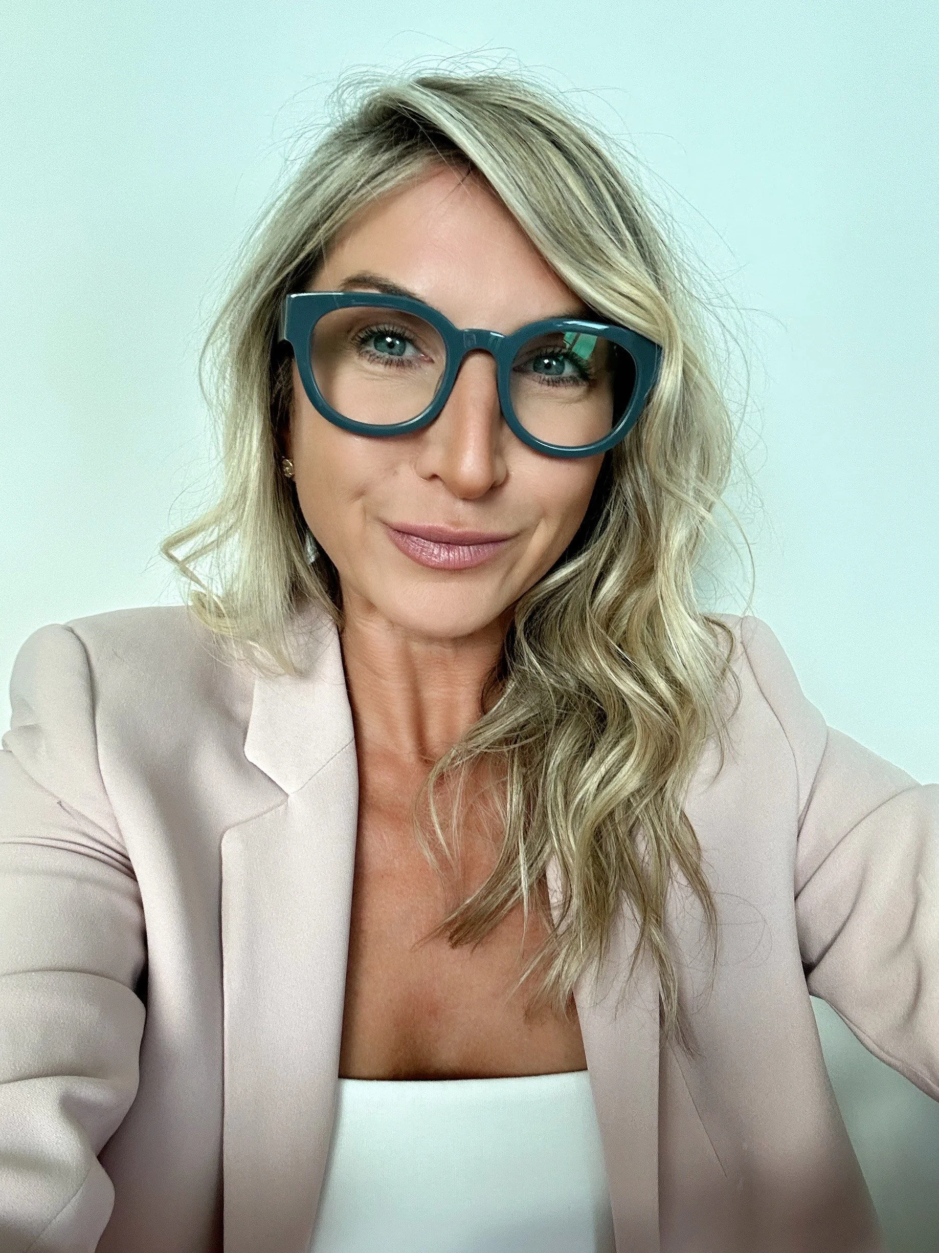 A woman with wavy blonde hair wearing large teal glasses, a beige blazer, and a white top, smiling softly in front of a plain light background.