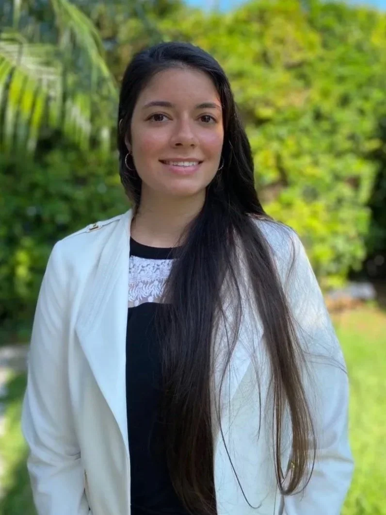 A young woman with long dark hair, wearing a white jacket and black top, standing outdoors in a garden with palm trees and green foliage under a blue sky.