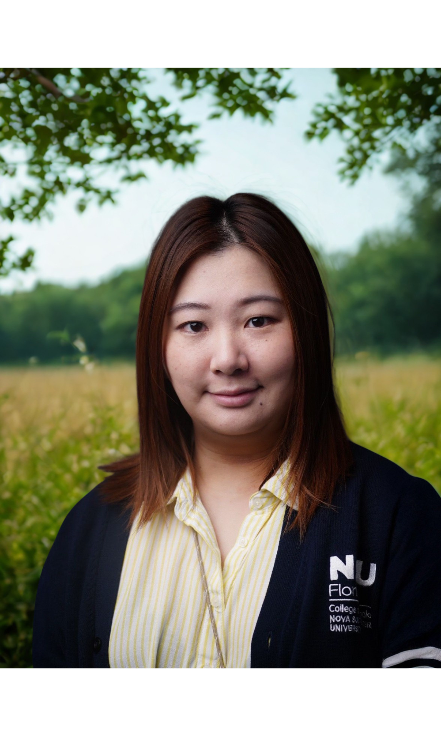 A portrait of a young woman with shoulder-length brown hair, wearing a striped yellow and white shirt and a black jacket with an embroidered logo on the chest, against a dark background.