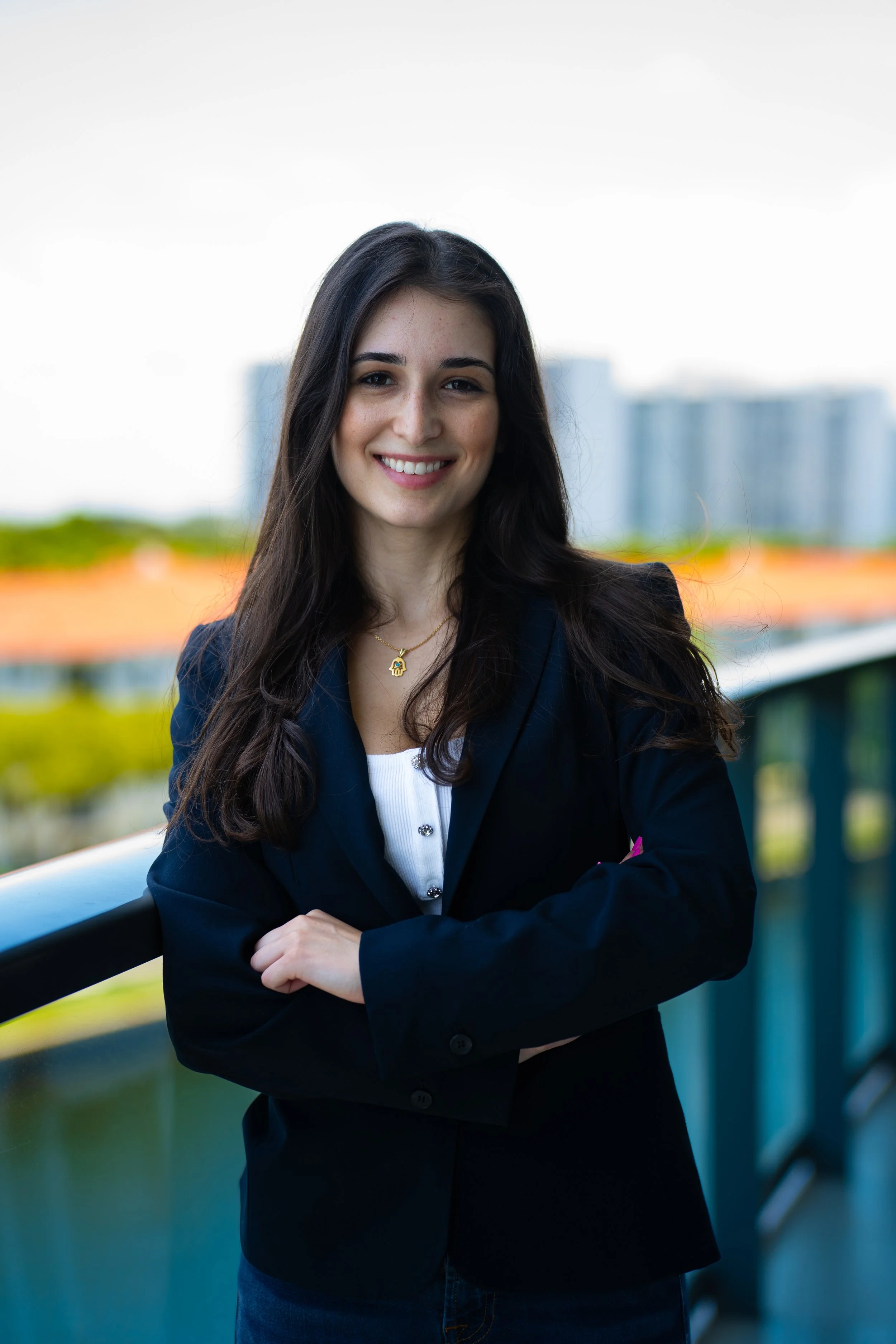 Young woman with long dark hair smiling, dressed in a dark blazer and white top, standing outdoors near a railing with buildings in the background.