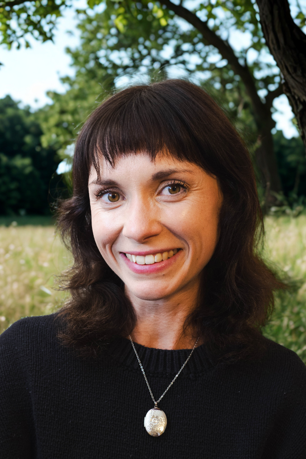 A woman with blonde hair in a bun, wearing dark makeup and earrings, smiling outdoors with green trees in the background.