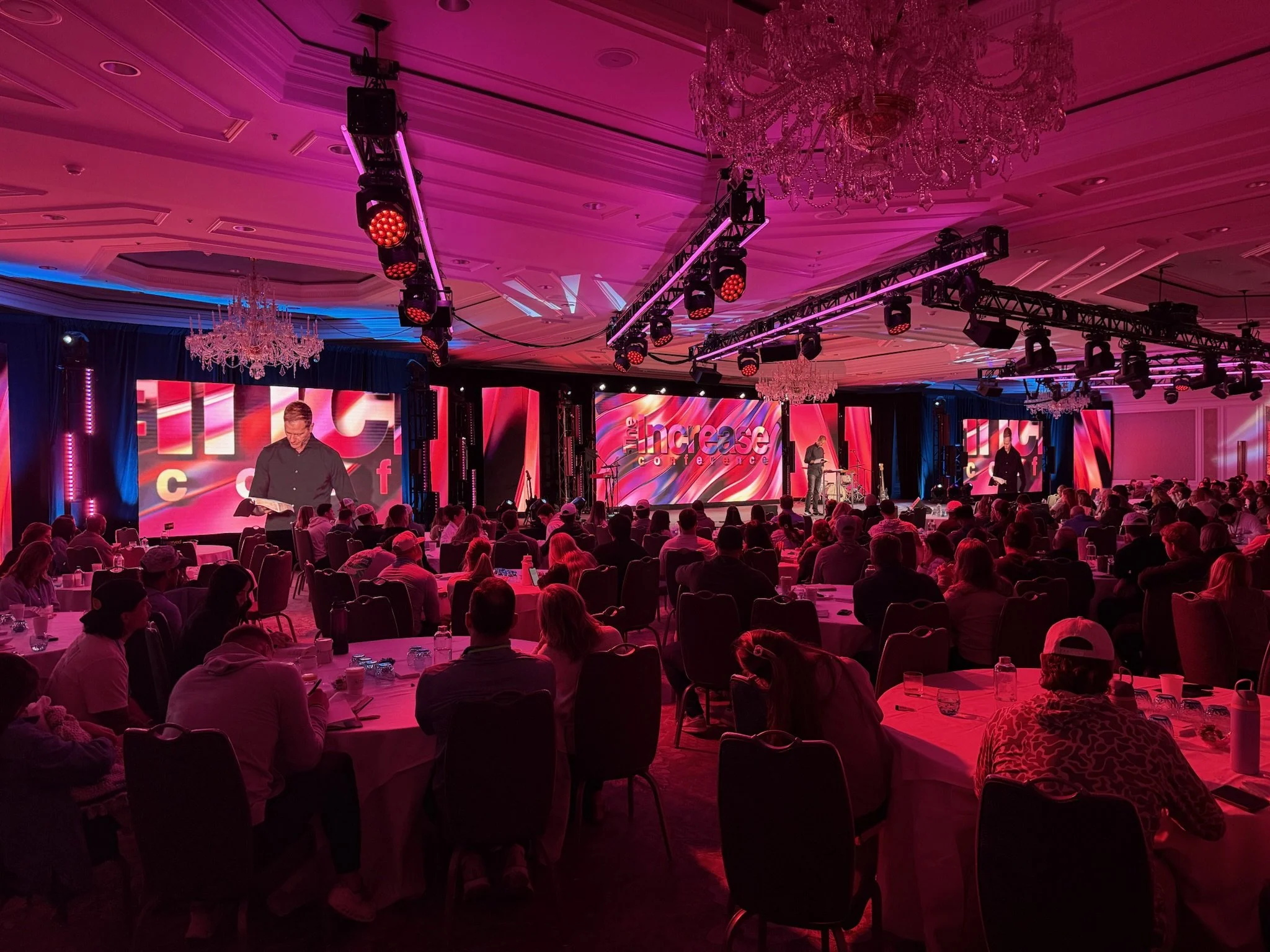 A conference hall with an audience seated at round tables, stage with multiple screens, lighting equipment, and chandeliers, during a presentation by a speaker.