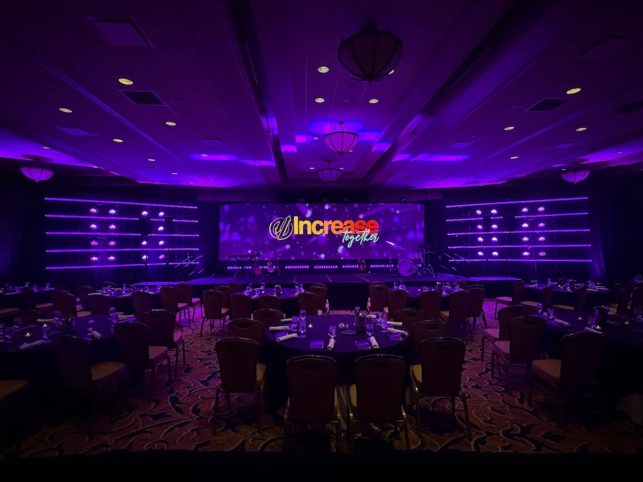 A large event space with purple lighting, round tables with chairs, and a stage with a screen displaying the words "Increase Together".
