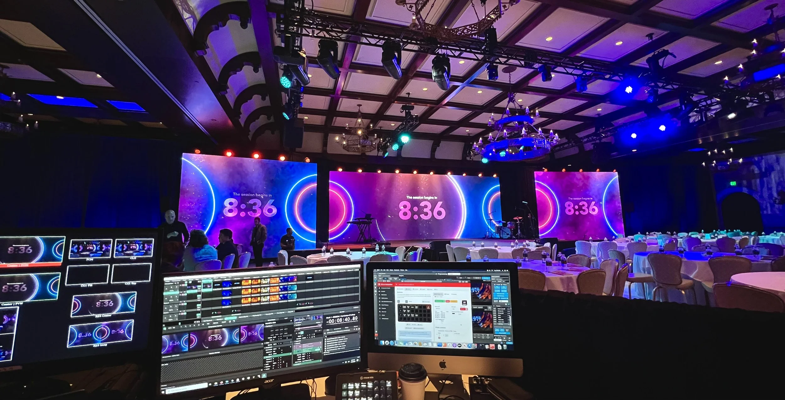 A large conference room or event hall with a stage and three big screens displaying the time 8:36. The room has round tables with chairs, set for an event. The ceiling has ornate woodwork, chandeliers, and numerous lighting fixtures. In the foreground, there is a control booth with multiple monitors and computers showing event controls and visuals.