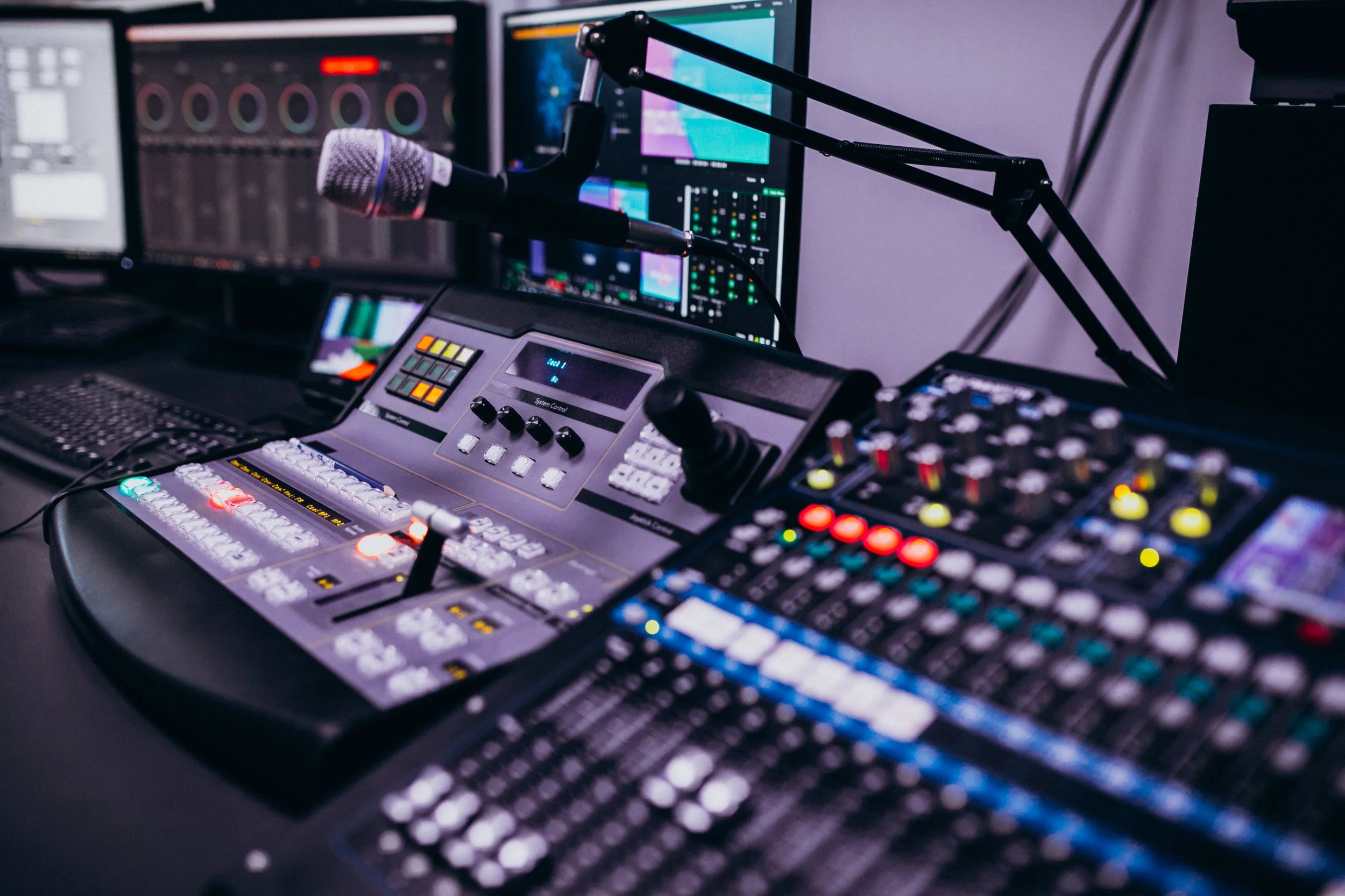 A professional radio broadcasting or sound mixing studio with multiple audio control panels, a microphone with a pop filter, and computer monitors displaying audio levels and editing software.