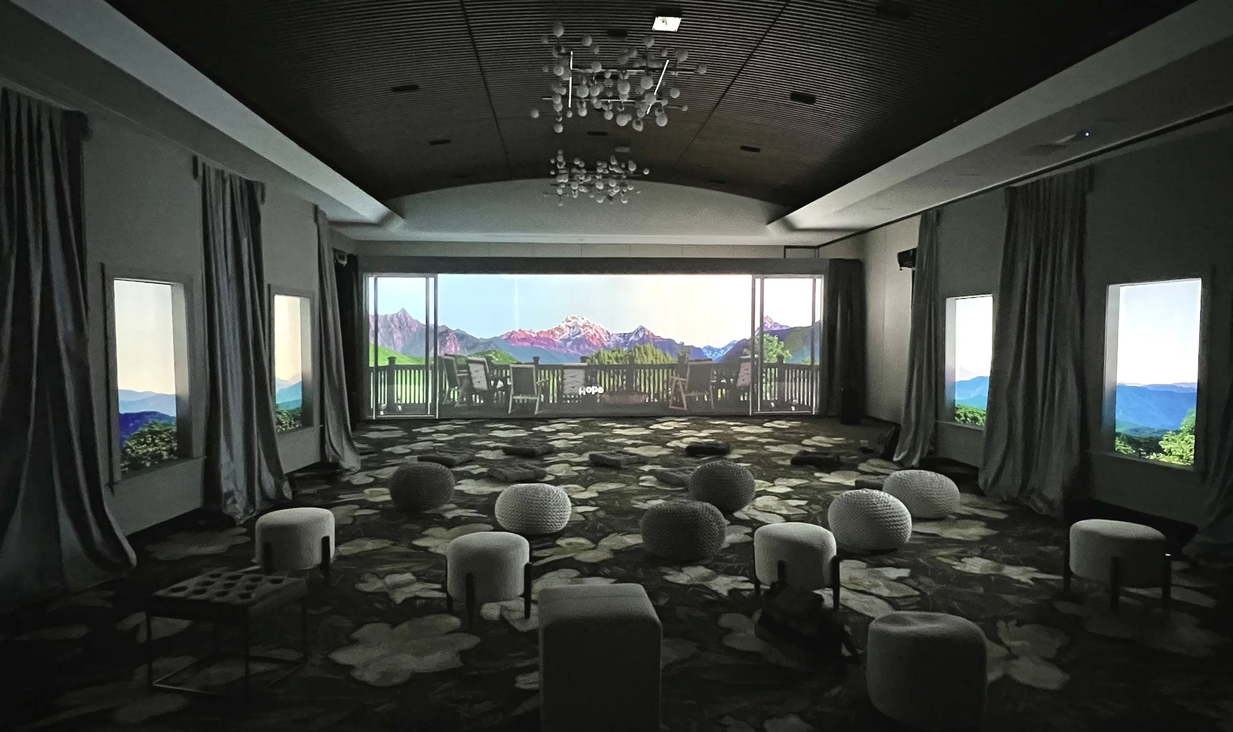 Inside a darkened room with large windows showcasing a mountain landscape, including snow-capped peaks, trees, and a blue sky. The room has a patterned carpet, white ottomans, and a balcony with chairs.