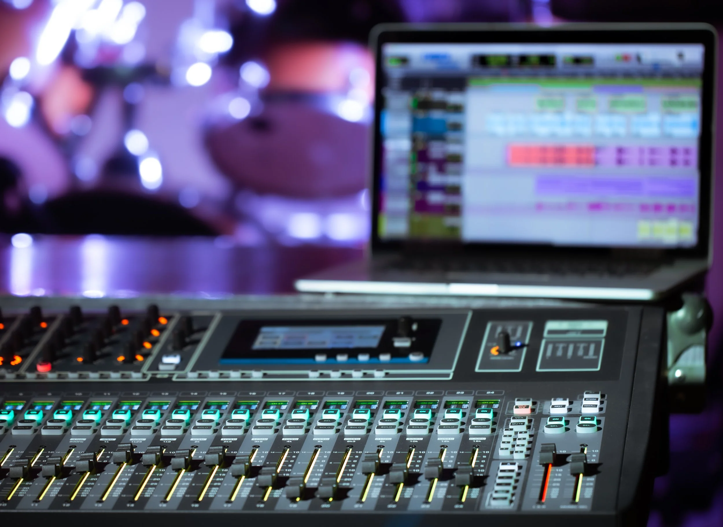 A music production mixing console with illuminated controls, monitor screen displaying digital audio workstation software, set in a dim, colorful environment with purple and blue lighting.