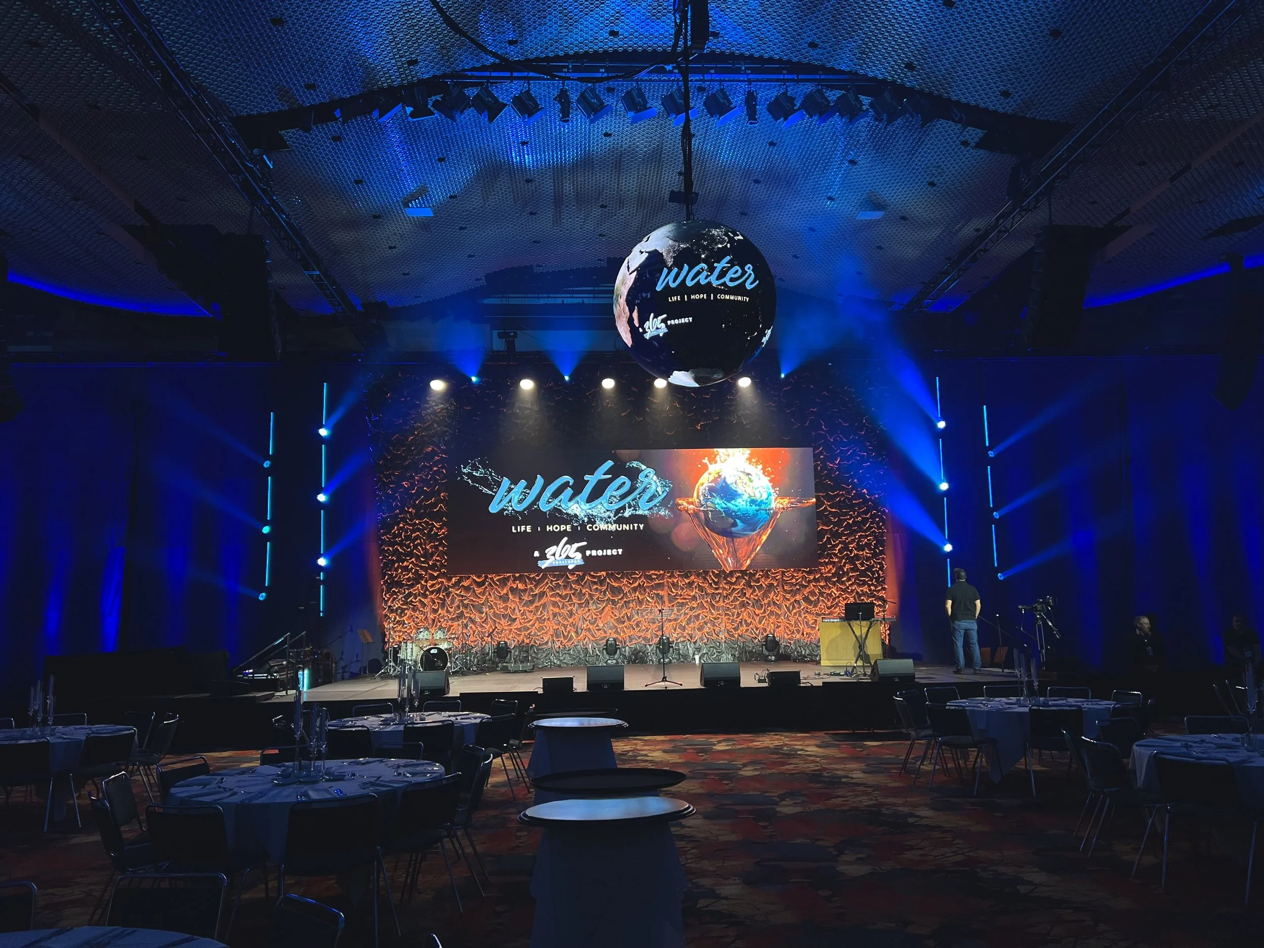 Concert stage with large LED screen displaying a globe and the words 'water, life, hope, community'. The stage is illuminated with blue and orange lights, with musical instruments and seating arrangements in the foreground, indicating preparations for a performance or event.