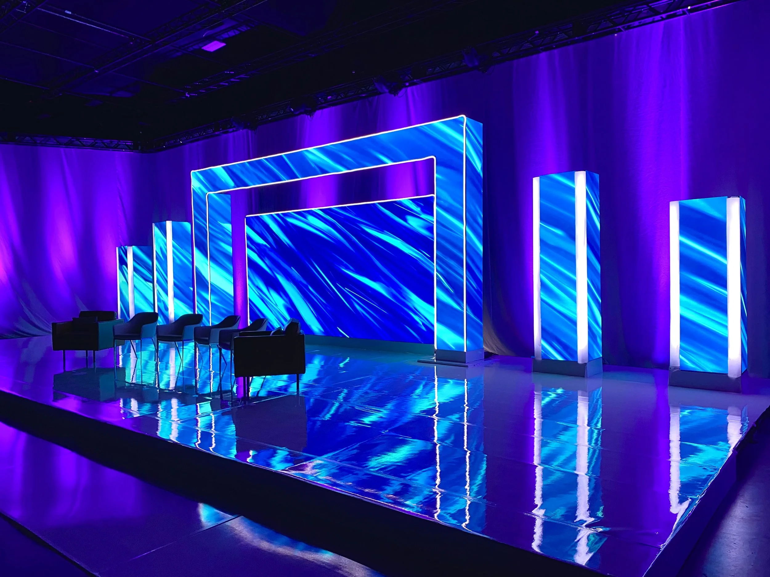 Stage with blue and purple lighting, electronic screens with blue patterns, chairs arranged in front, and a purple backdrop.