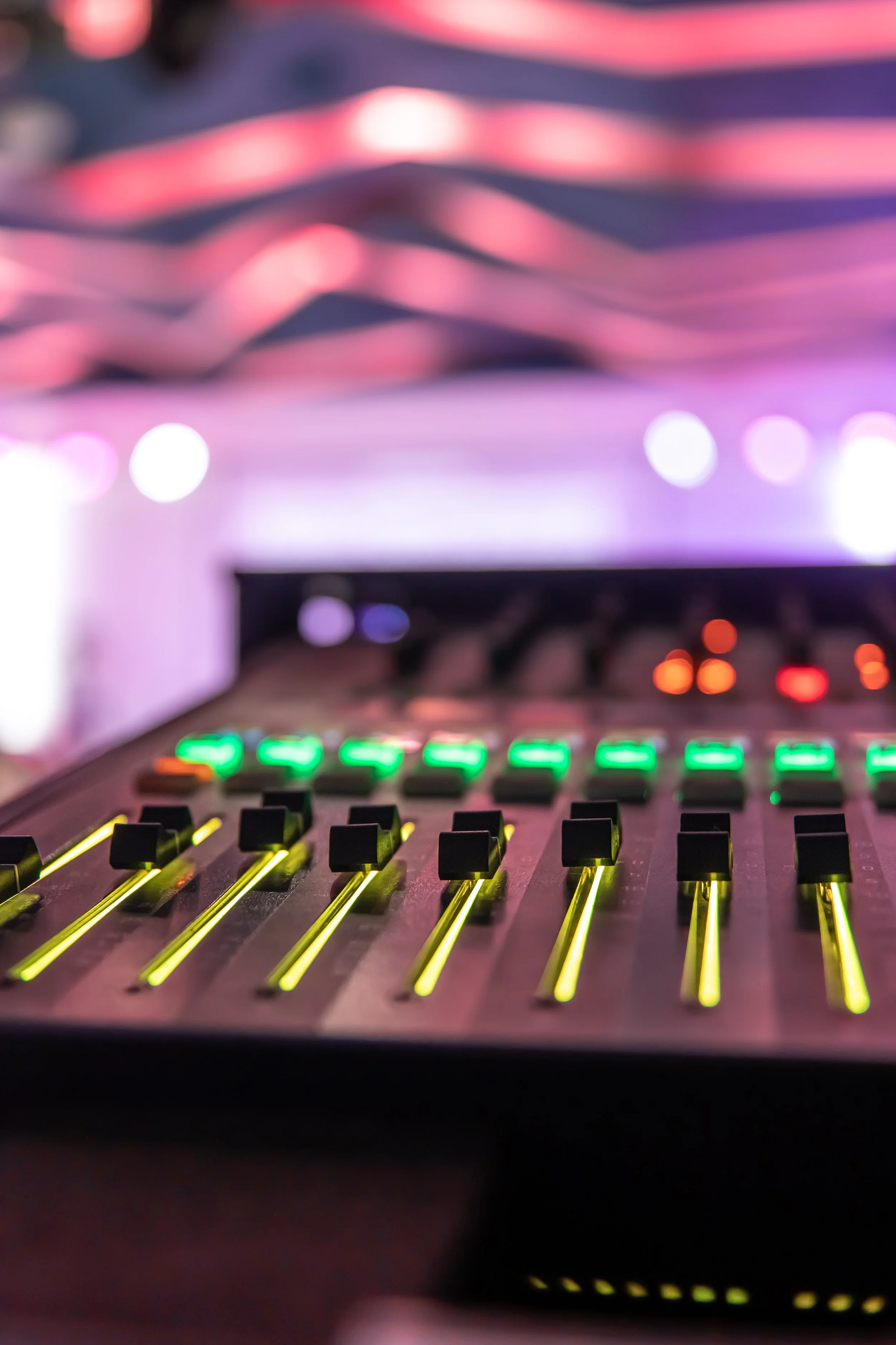 Close-up of a DJ mixing console with colorful illuminated sliders and controls, blurred background with screens displaying abstract visuals.
