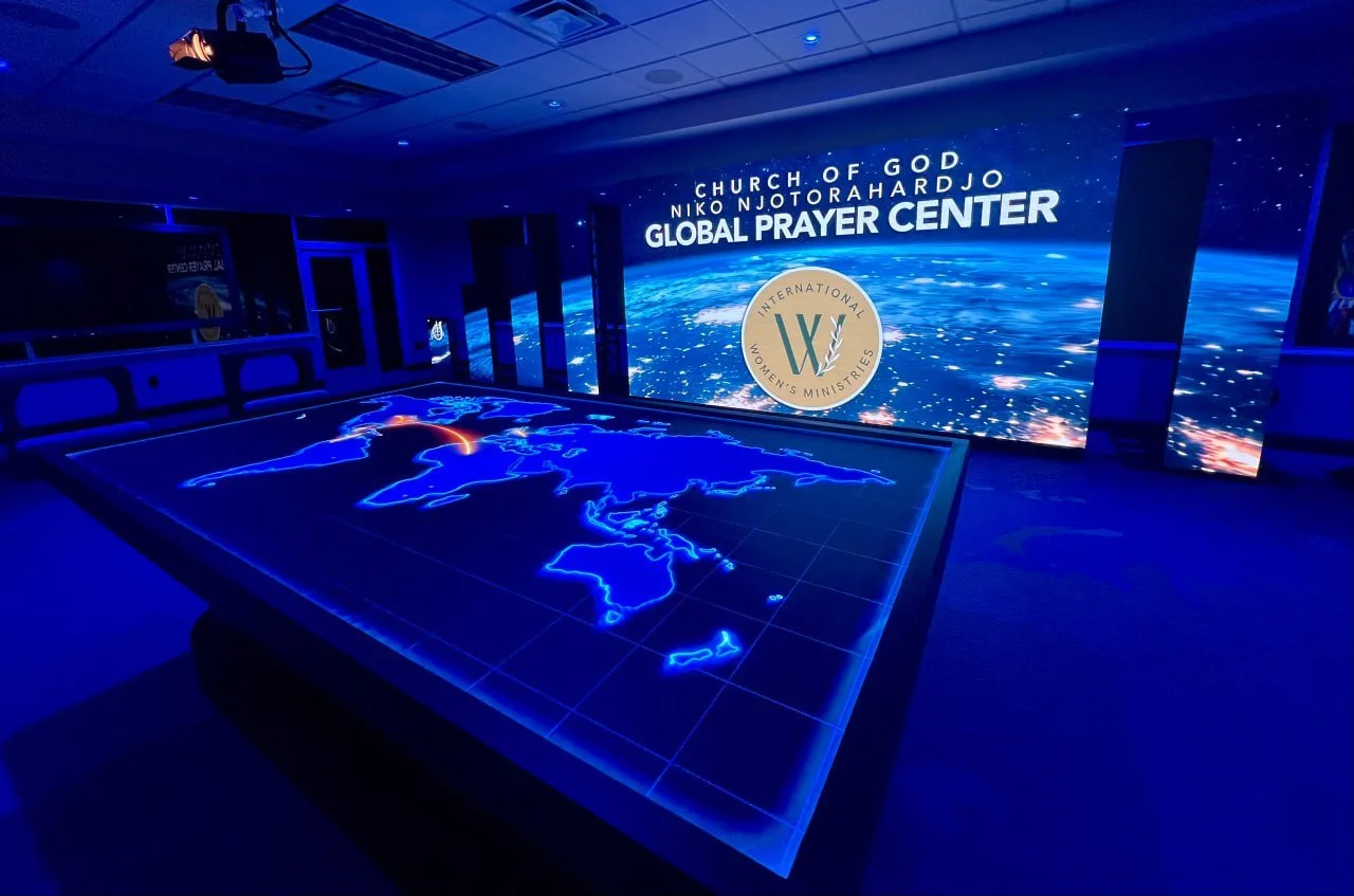 A large digital screen displaying Earth from space with the text 'Church of God Niko NjoroHardjo Global Prayer Center' and the logo of International Women's Ministries. A 3D interactive map of the world is on a table in the foreground, showing illuminated outlines of continents with red lines connecting different locations.
