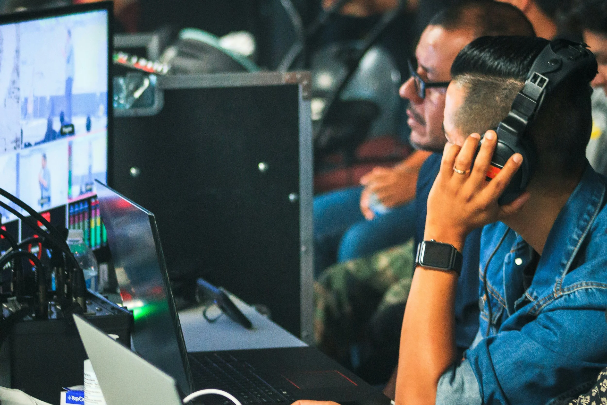 Two people working at computers in a tech or gaming environment, one wearing headphones, with various electronic equipment around them.