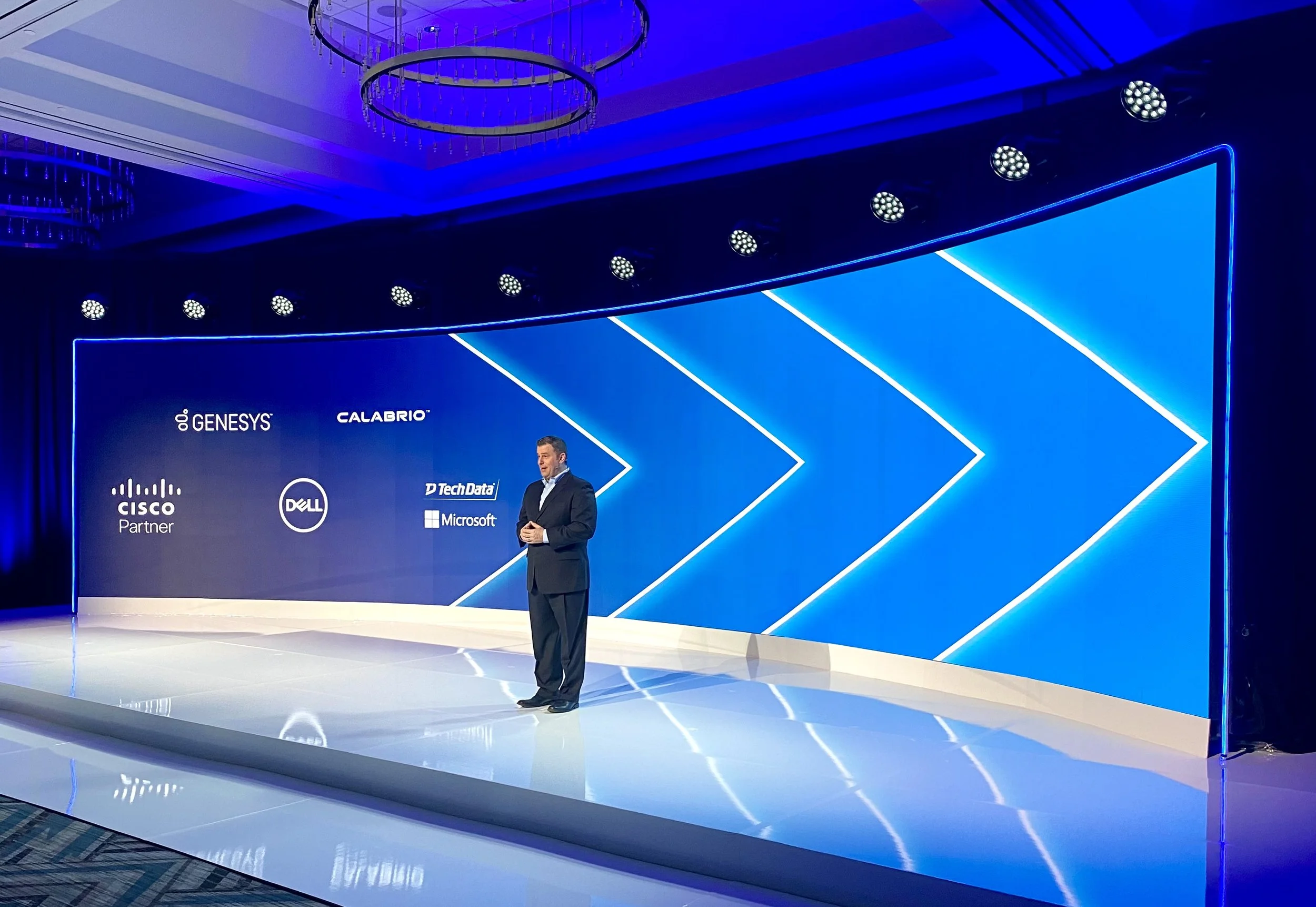 A man standing on a stage at a tech conference with a blue geometric background displaying logos of companies such as Genesys, Calabrio, Cisco, Dell, Microsoft, and Tech Data. The stage is illuminated with blue lighting and a circular chandelier hangs from the ceiling.