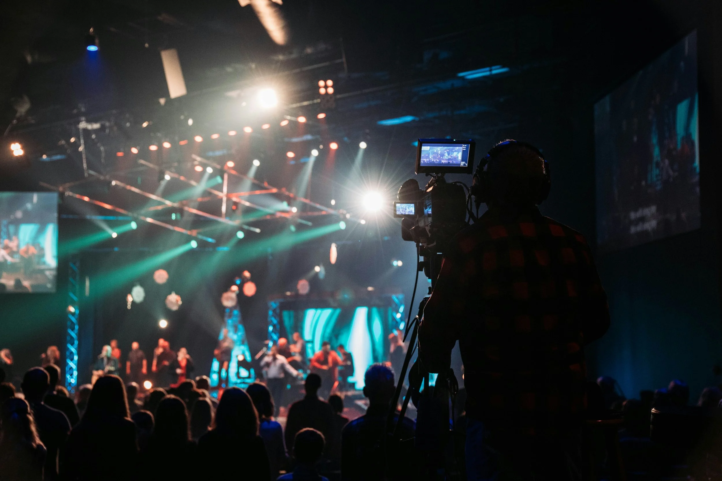 A concert scene with an audience watching a stage performance, colorful lighting, and a camera operator recording the event.