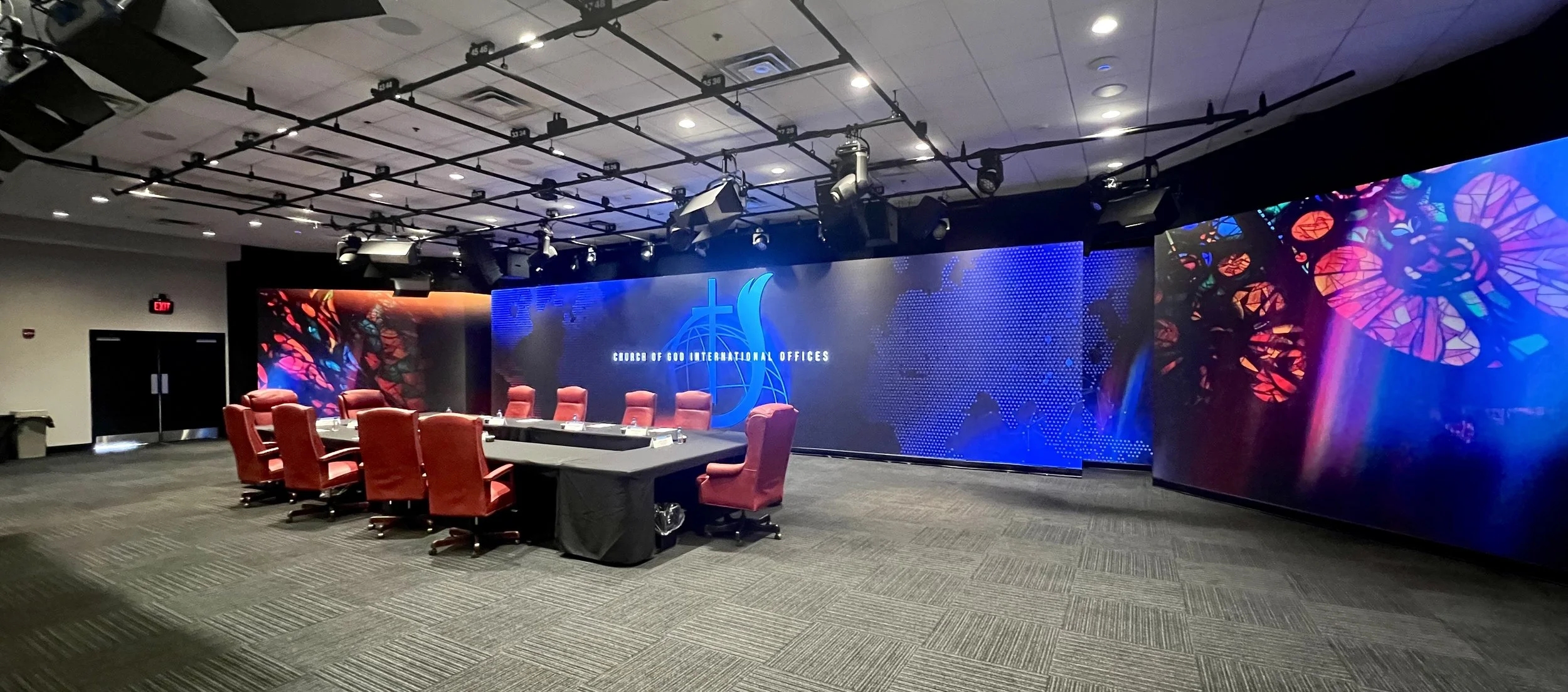 Conference room with a large digital display wall, red office chairs around a black table, and colorful stained glass imagery on the display, with the words 'Church of God International Offices'.