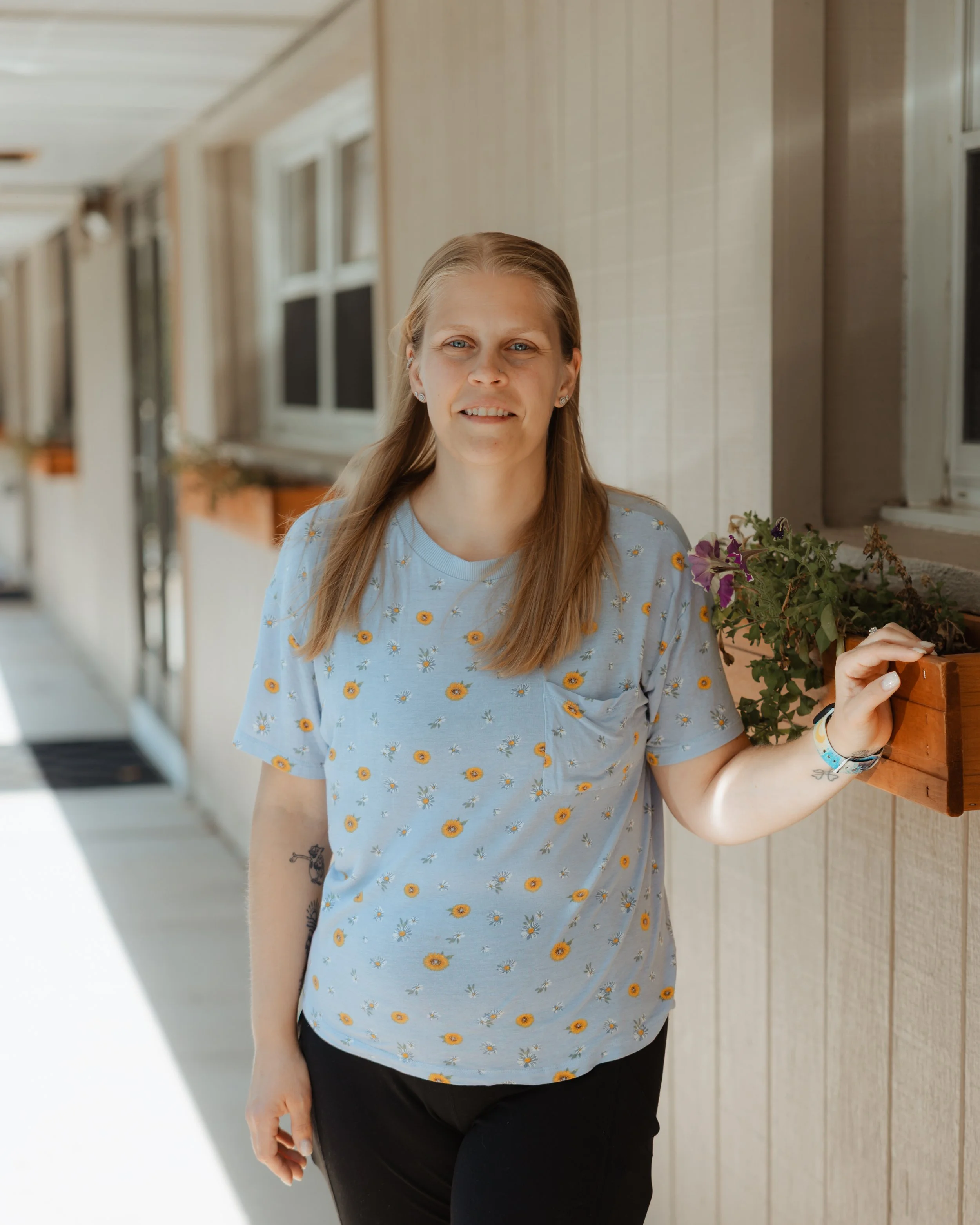 A woman with blonde hair wearing a light blue shirt with a daisy pattern, standing outdoors beside a wooden wall with windows, smiling at the camera.