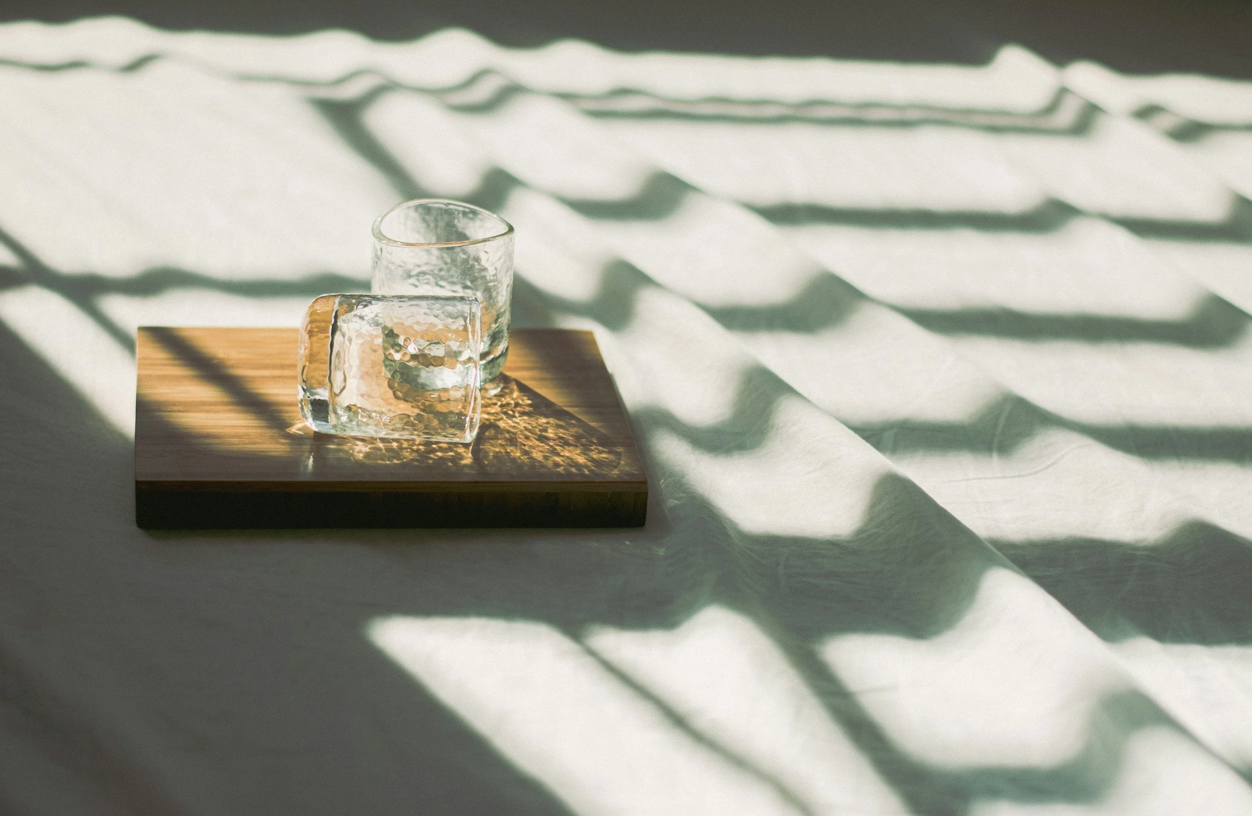 Two textured glass cups sit on a wooden tray with shadows cast on a light-colored fabric.