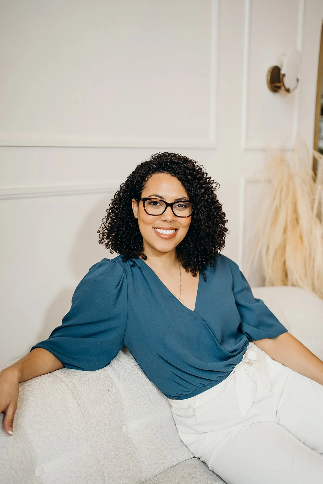 A woman with curly black hair and glasses smiling while sitting on a light-colored sofa in a cream-colored room.