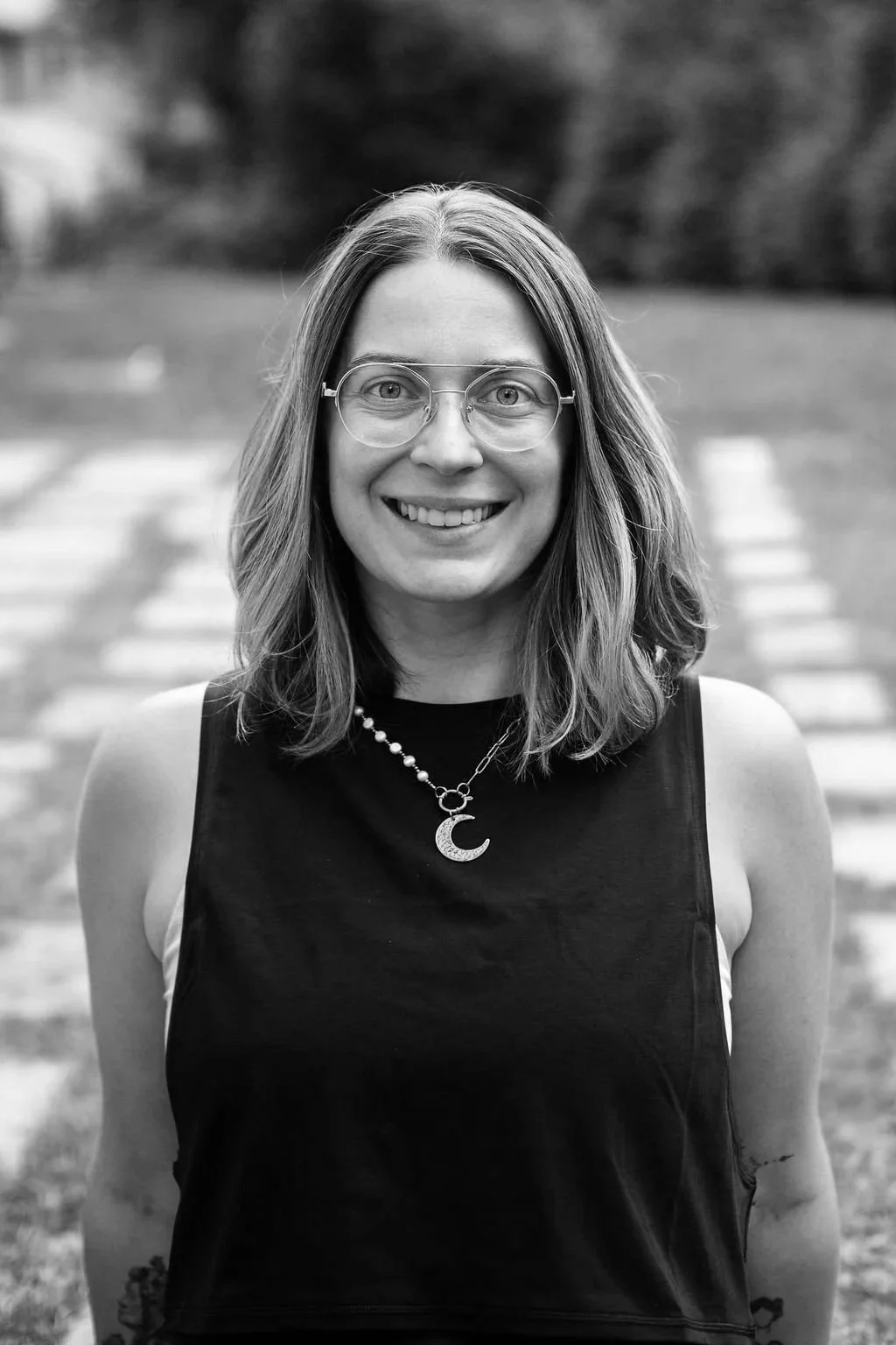 A woman with wavy hair, wearing glasses, a baseball cap, and a light-colored sweater, is looking at the camera, in black and white.