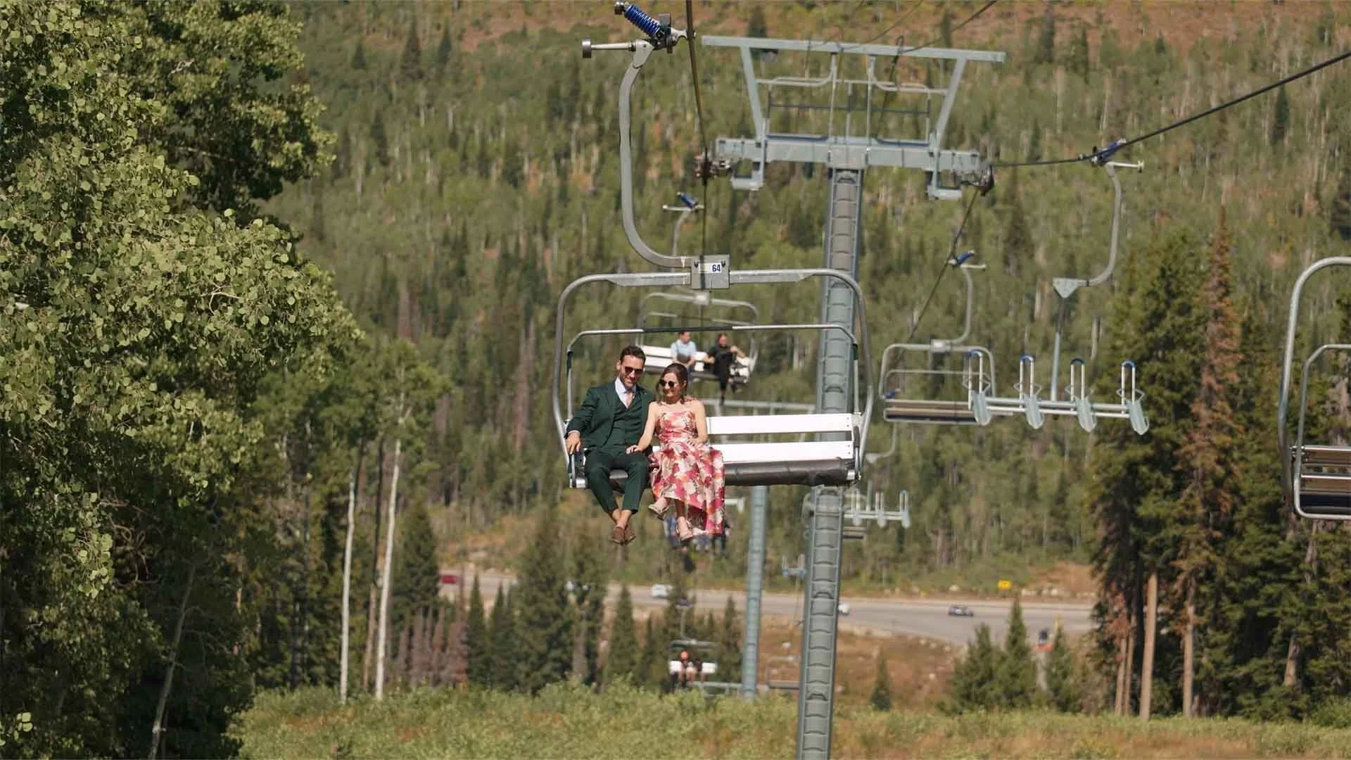 Couple riding a ski lift in a mountainous, forested area during daytime.
