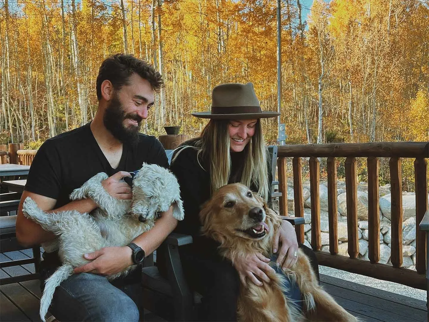 A man and woman sitting on a wooden deck with autumn trees in the background, each holding a dog and smiling.