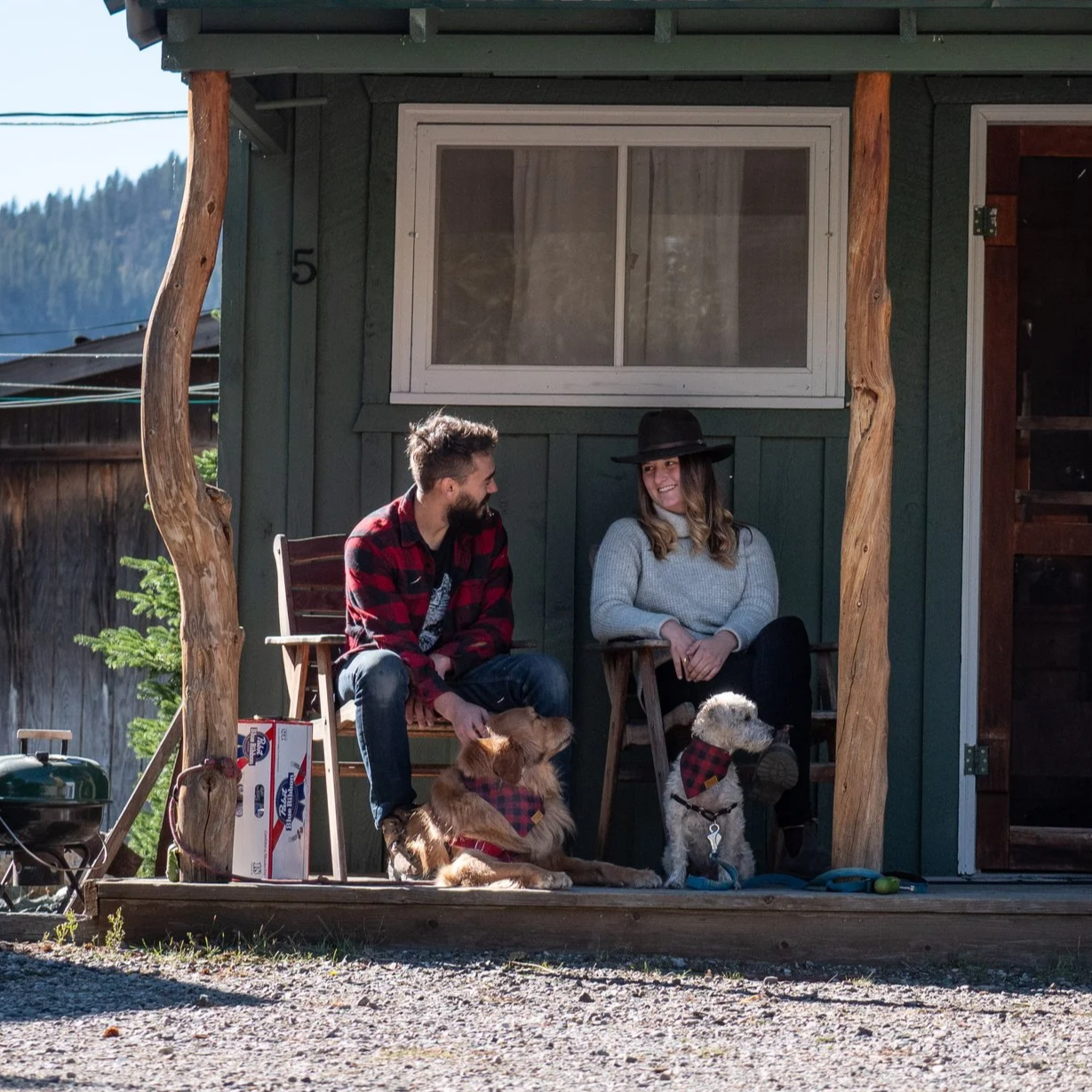 A man and woman sitting on the porch of a cabin, smiling at each other, with two dogs seated at their feet. The man is wearing a red and black plaid shirt, and the woman is wearing a gray sweater and a black hat. There is a box of Budweiser beer and a green barbecue grill on the porch.