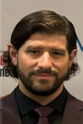 A man with dark hair and a beard wearing a dark suit and tie, standing indoors with a neutral background.