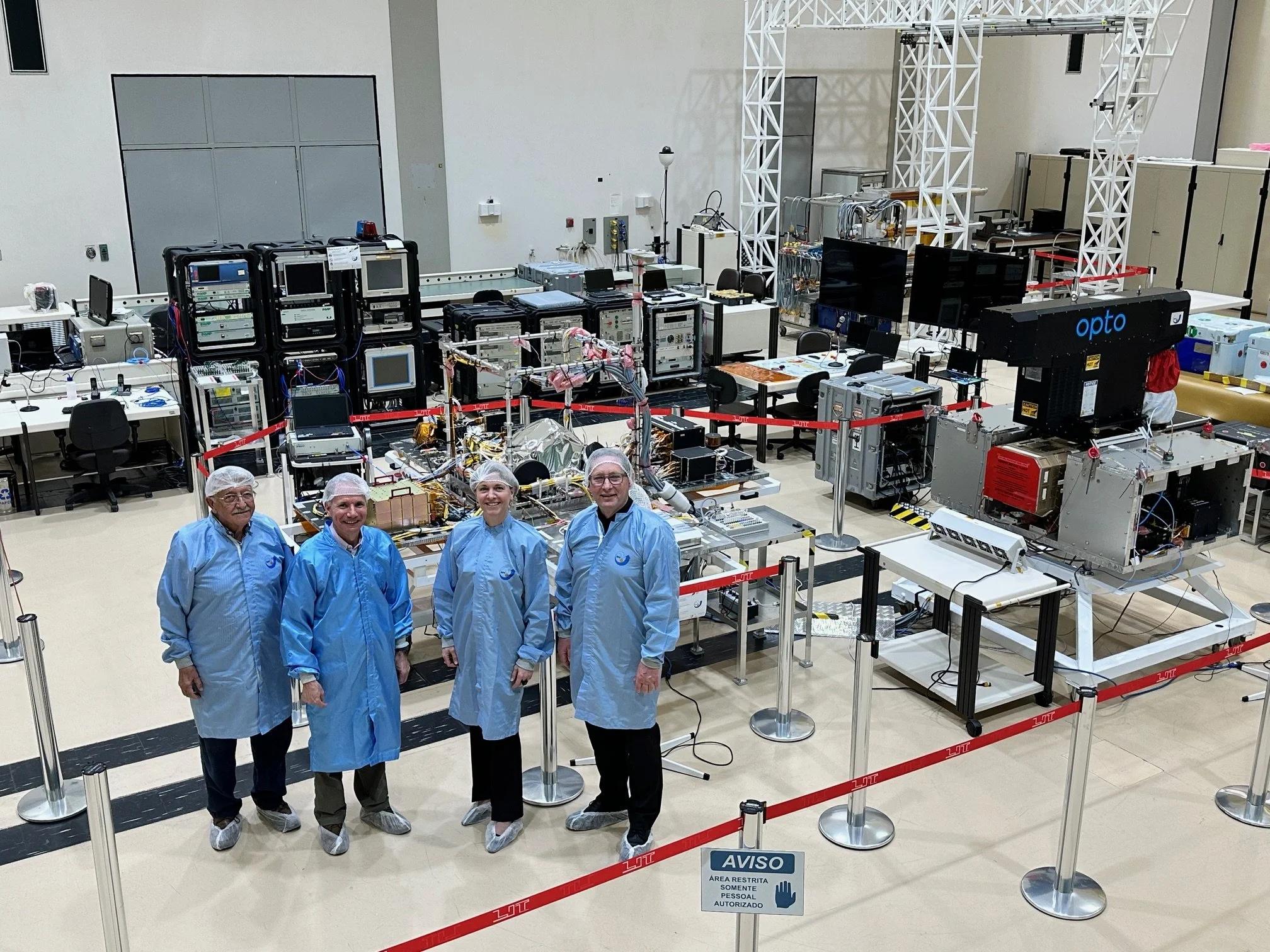 Four people in lab coats and hairnets standing in front of complex scientific equipment in a laboratory setting.
