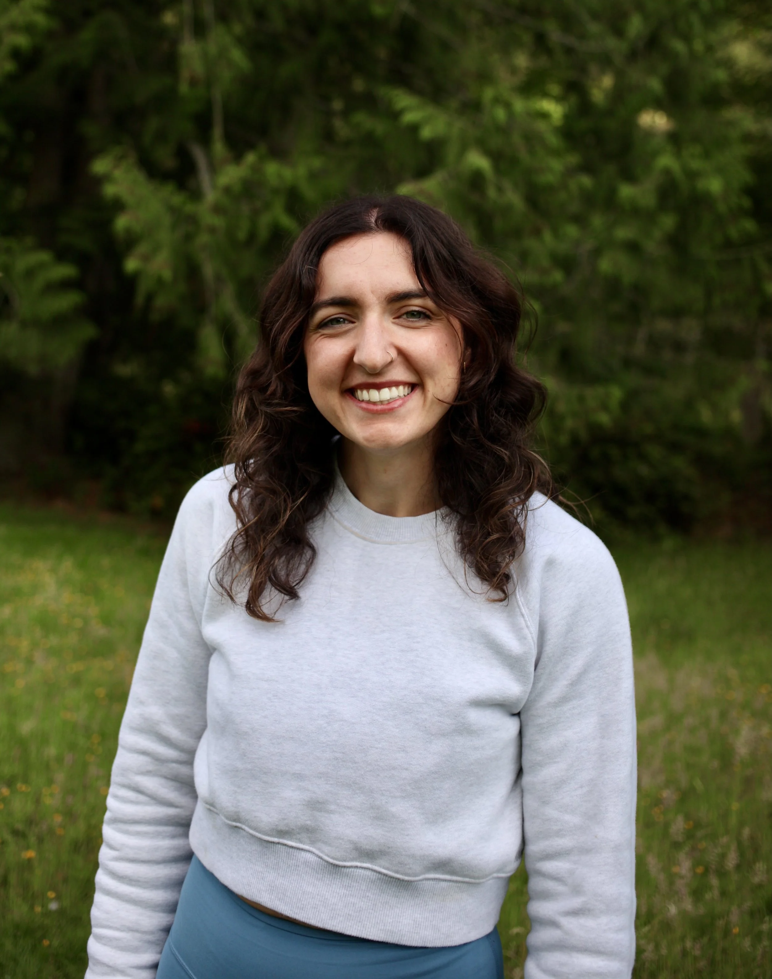 A young woman with dark, wavy hair, smiling outdoors in a green, wooded area, wearing a light gray sweatshirt and blue pants.