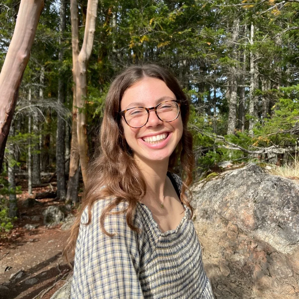 A young woman with long curly hair and glasses smiling in a forested area.