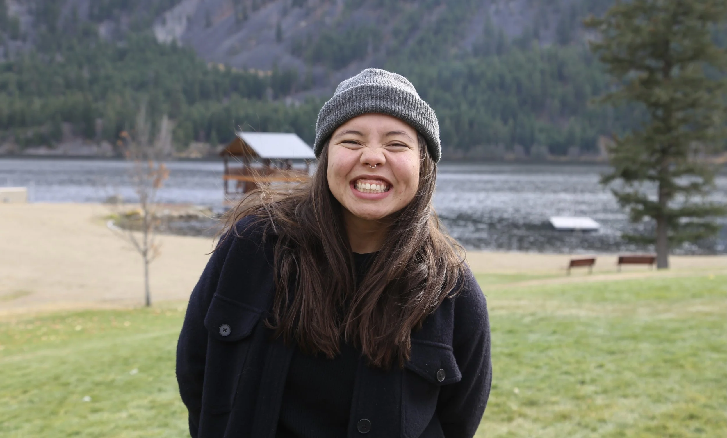 Young woman smiling with teeth showing, wearing a gray knit beanie and a dark jacket, standing outdoors near a lake with mountains and trees in the background.