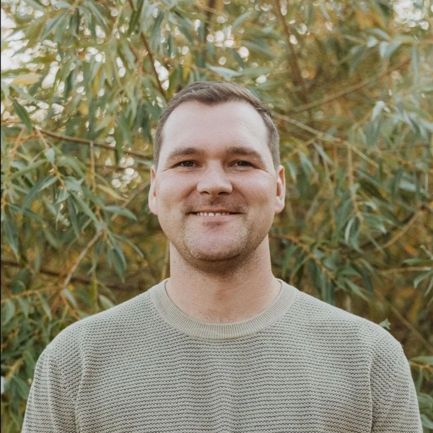 A man with short dark hair and a light beard smiling outdoors, wearing a light gray textured sweater, with green leafy bushes in the background.