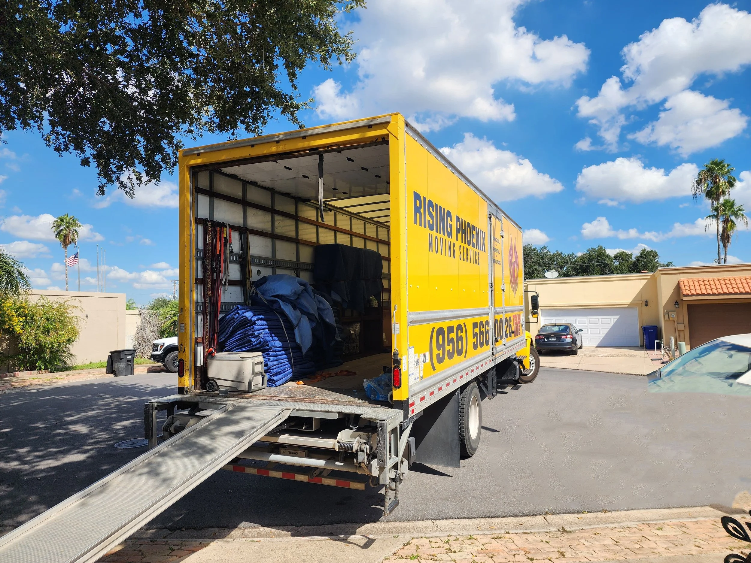 Open yellow moving truck labeled 'Rising Phoenix Moving Service' parked in a residential neighborhood with a blue sky and trees in the background.