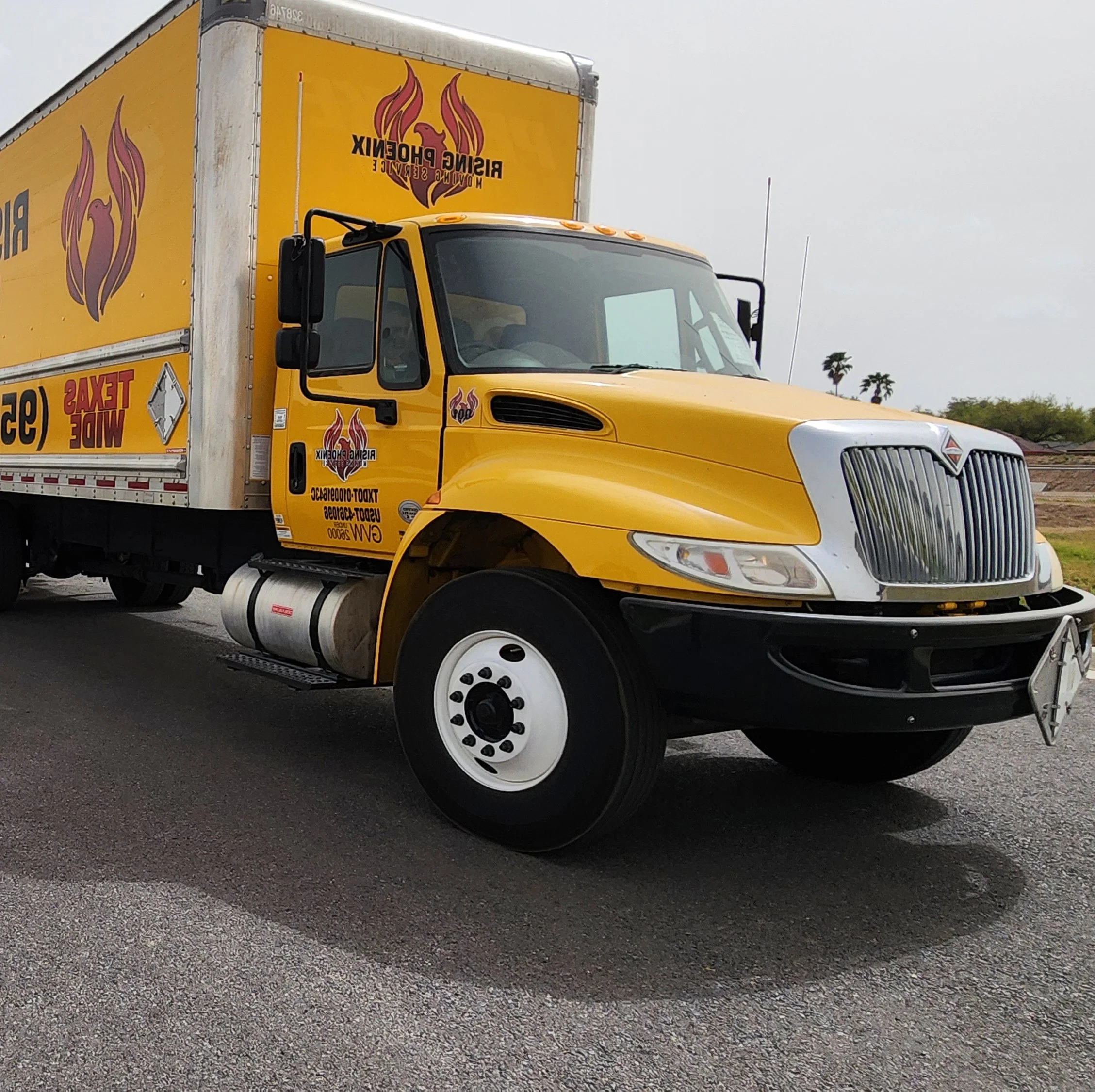 Yellow delivery truck with logo of a phoenix and text 'Rising Phoenix Moving Service' on the side, parked on a road with palm trees in the background under an overcast sky.